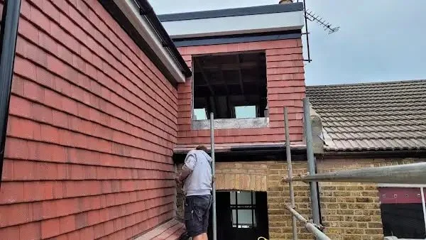 A man working on the exterior of a brick house under construction or renovation, with scaffolding next to him and a partially open window above.