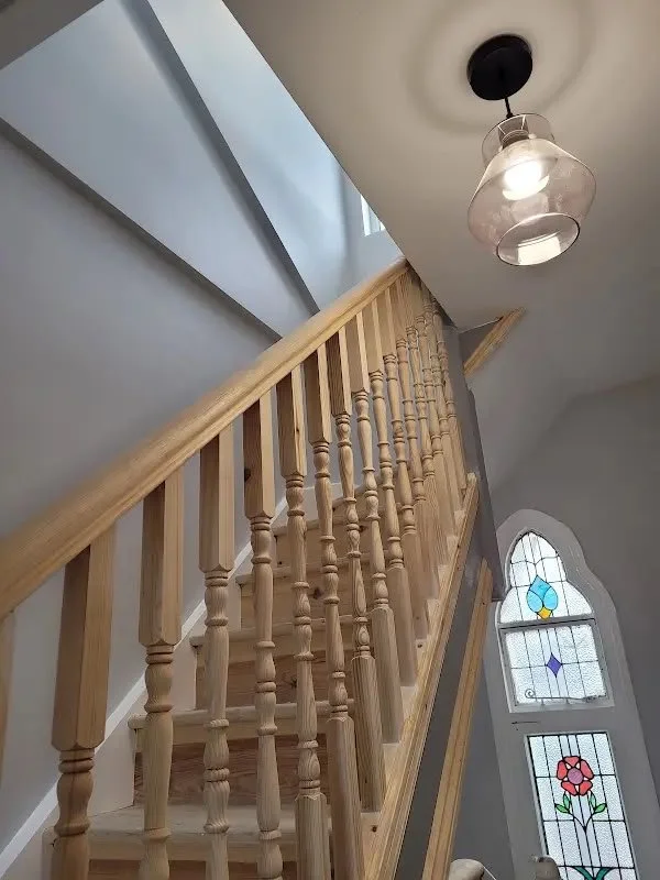 Wooden staircase with spindles and handrail, gray walls, stained glass window with floral and geometric design, hanging light fixture with glass shade.
