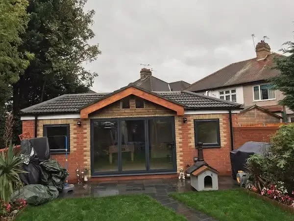 Backyard view of a brick house with black sliding doors, a small roof extension, and a landscaped garden with grass, flowers, trees, and outdoor items, including a doghouse and covered equipment.