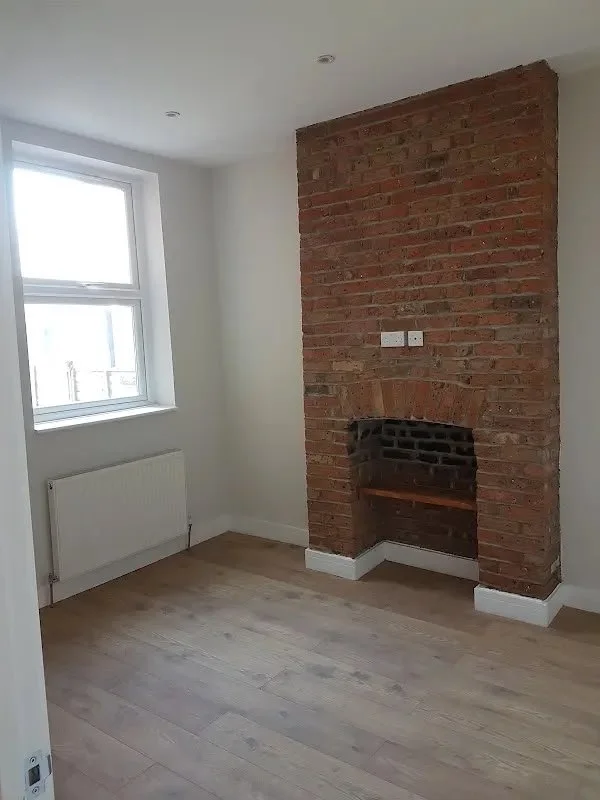 Empty room with white walls, wood flooring, a large window, and a brick fireplace without a mantle.