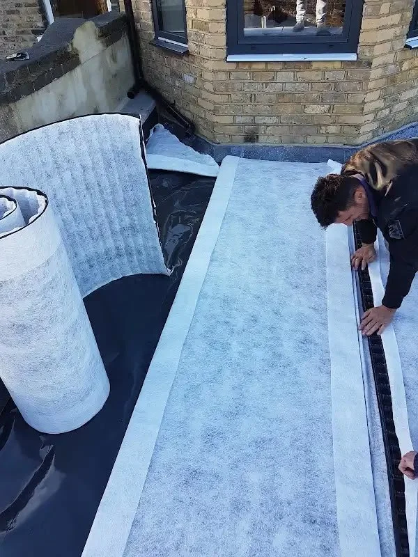 A man installing a flat roof waterproofing membrane with a ruler on a rooftop. Rolls of waterproofing material are nearby, next to a brick building with windows.