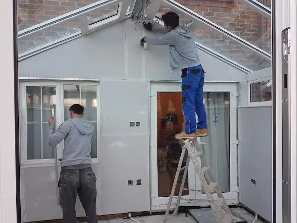 Two workers are installing or repairing a glass ceiling in a sunroom using a ladder and tools.
