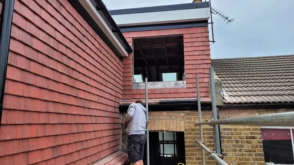A construction worker in gray sweatshirt and shorts working on the exterior of a brick house, with scaffolding surrounding the building. The house has red brick walls, and part of the upper story is under construction with open window spaces and roofing work in progress.