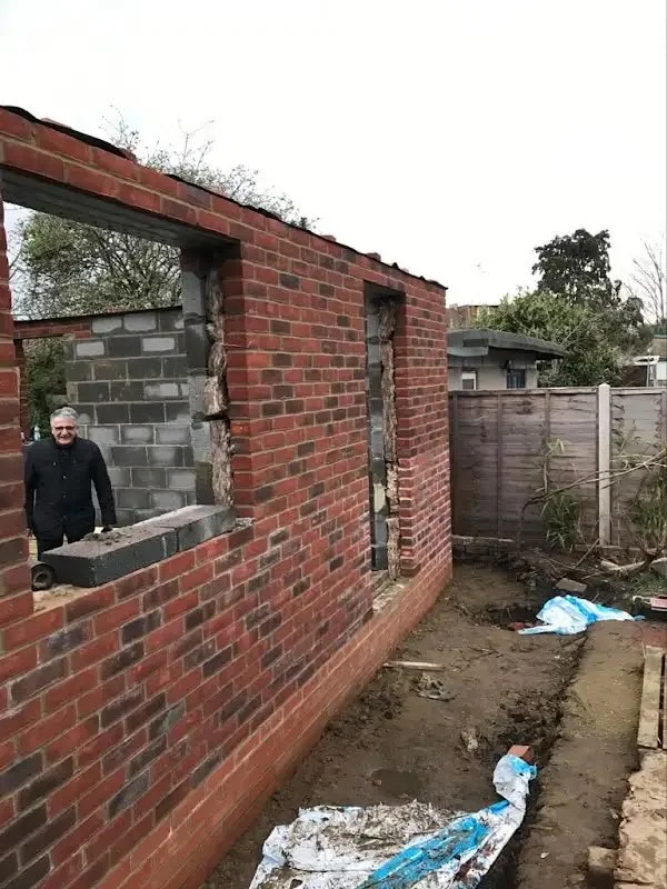 A man standing inside a brick building under construction with some window openings, in a backyard with a wooden fence and trees in the background.