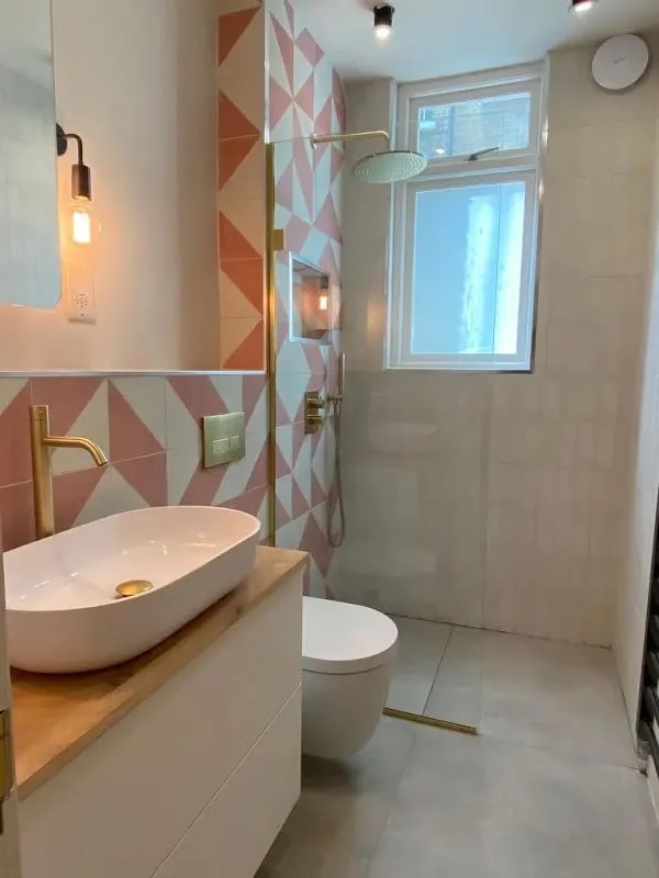 Modern bathroom with geometric patterned wall, white vessel sink with gold faucet, wall-mounted mirror, toilet, and walk-in shower with rain showerhead near a window.