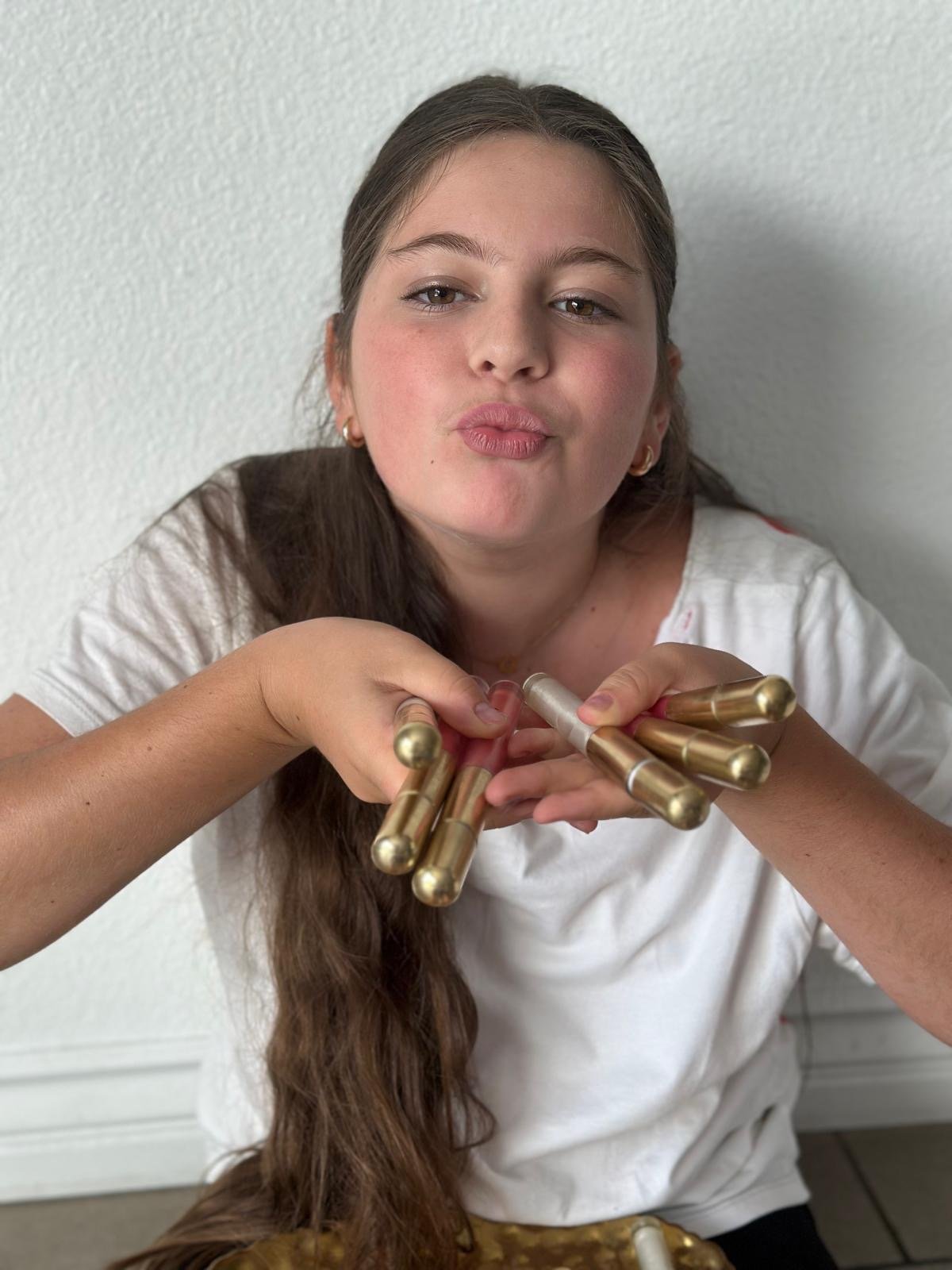 A young woman with brown hair, wearing a white top and small hoop earrings, making a duck face while holding several lip gloss tubes in her hands against a plain white wall.