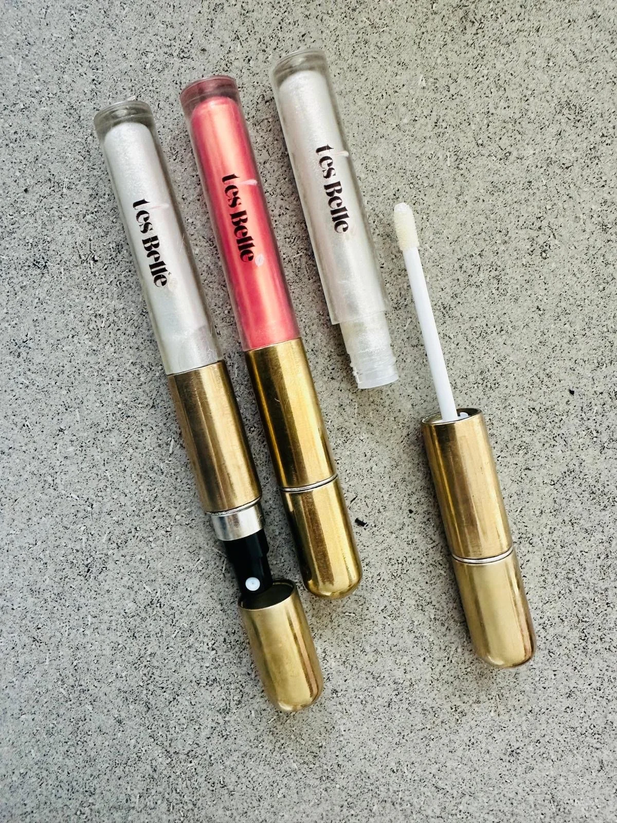 Four lip gloss tubes with gold caps on a textured surface, one tube is open with a white applicator brush resting on the cap.