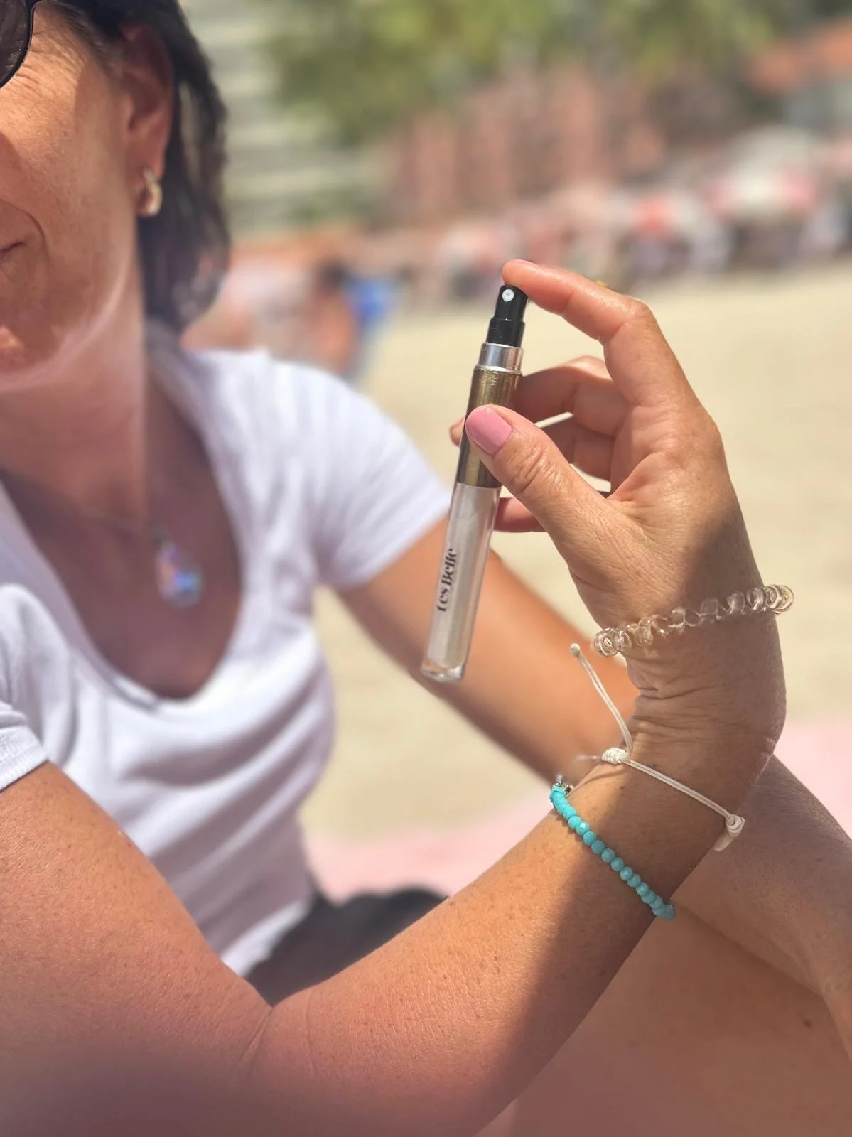 A woman holding a test tube at an outdoor setting. She is wearing a white shirt, bracelets, and a necklace, with a blurred background of outdoor scene.
