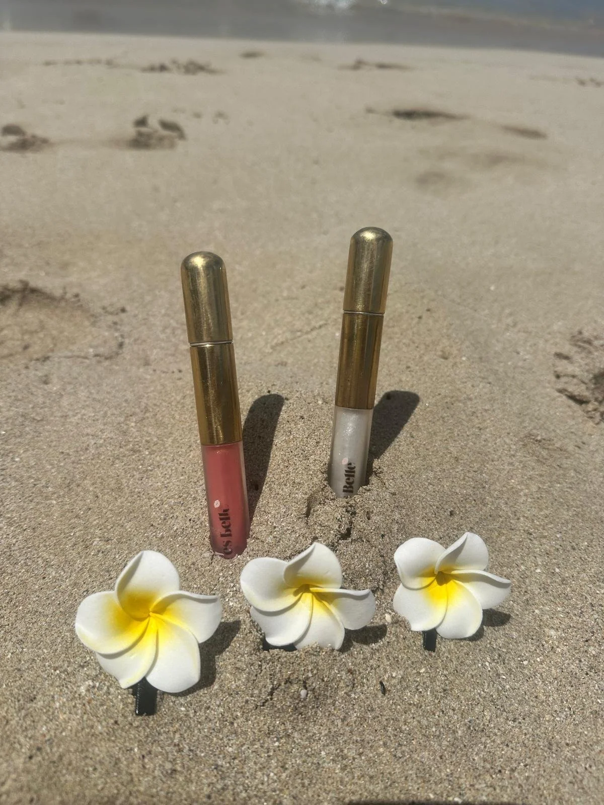 Two lip gloss tubes partially buried in the sand on a beach, surrounded by three white and yellow plumeria flowers.