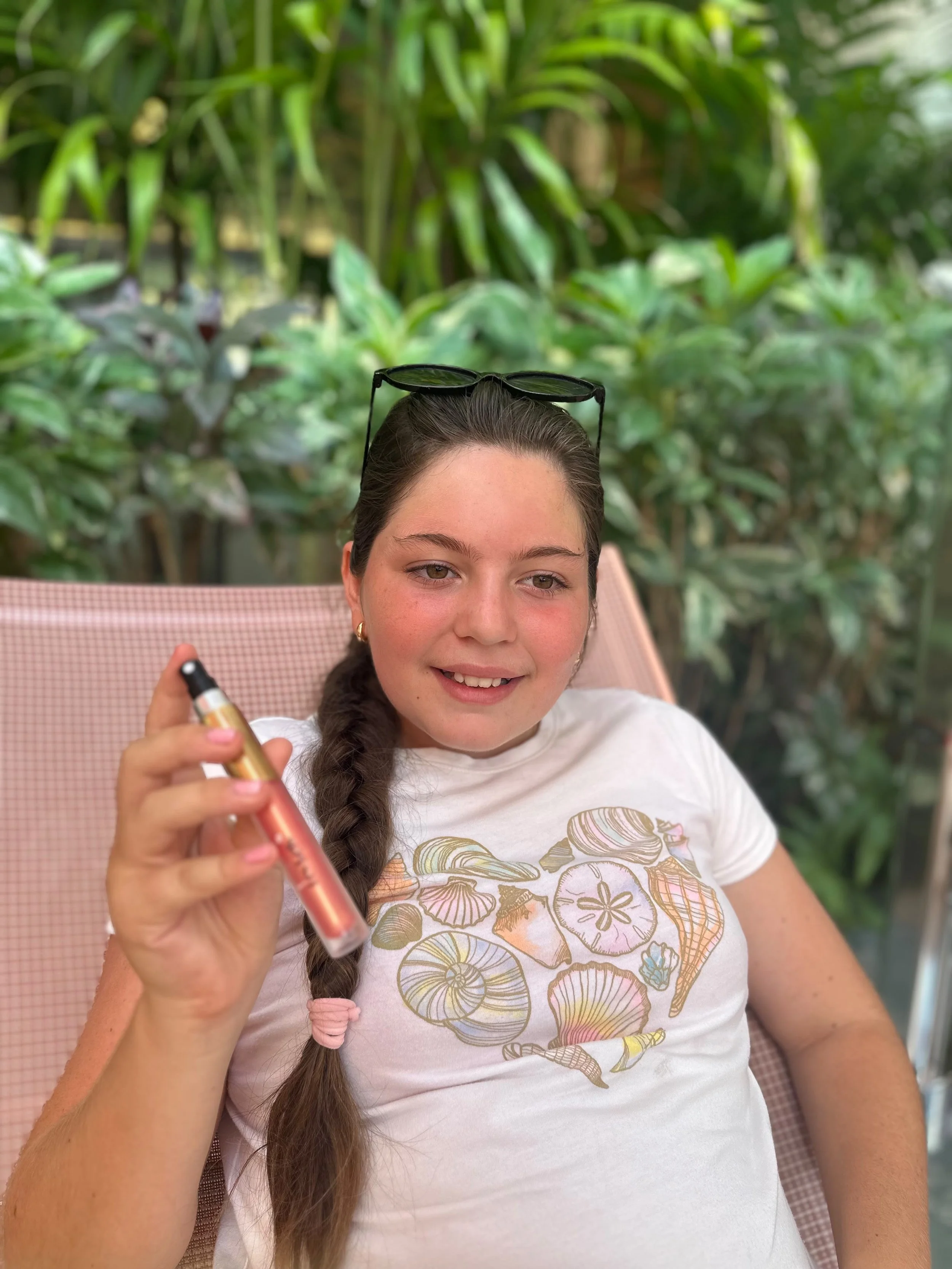 A young woman with long braided hair, wearing a white t-shirt with a colorful seashell print, is sitting outdoors and smiling while holding a vape pen. She has sunglasses on her head and is sitting in front of lush green plants.