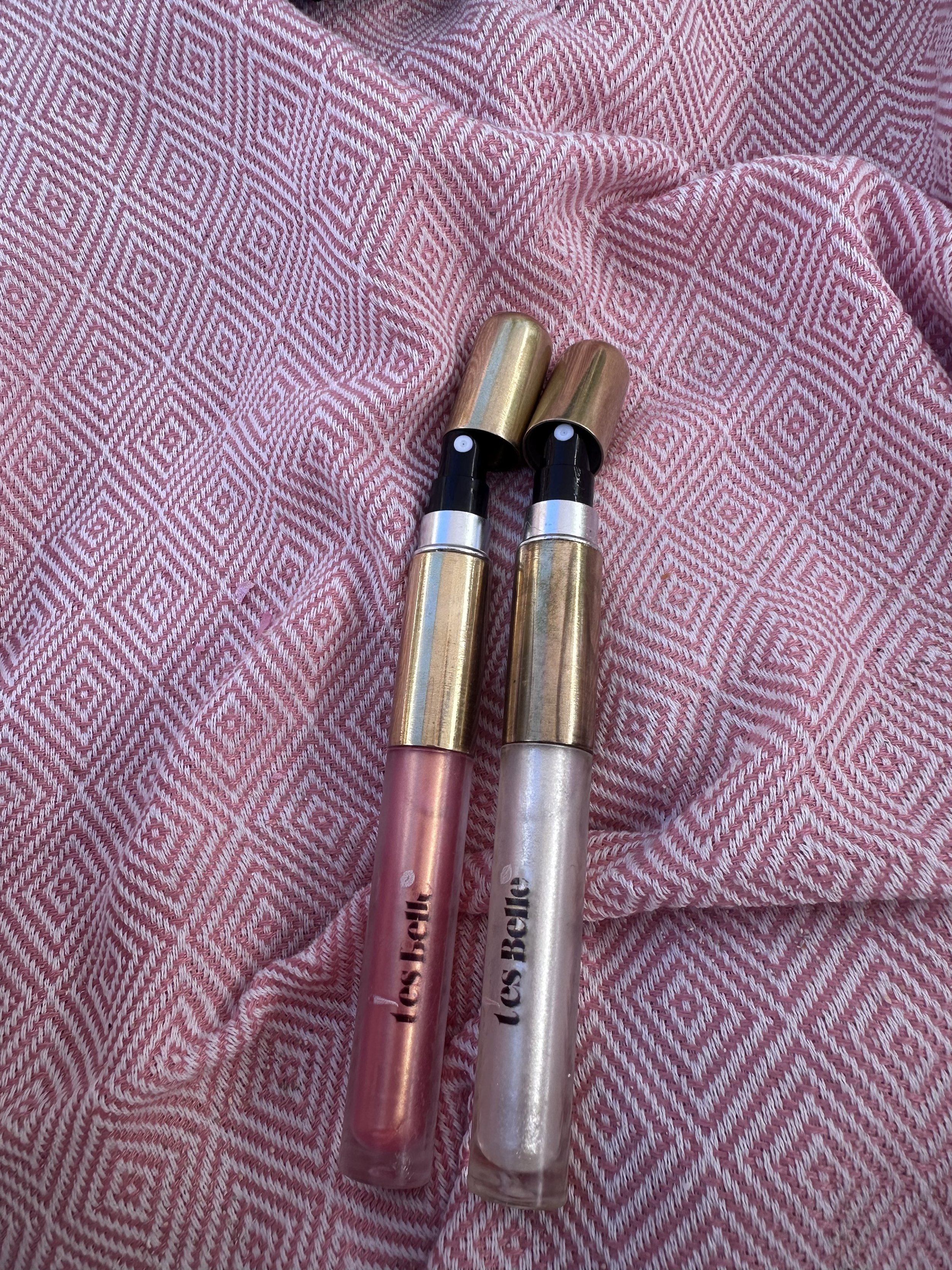 Two lip gloss tubes with gold caps and pink and white bodies, placed on a pink patterned fabric background.