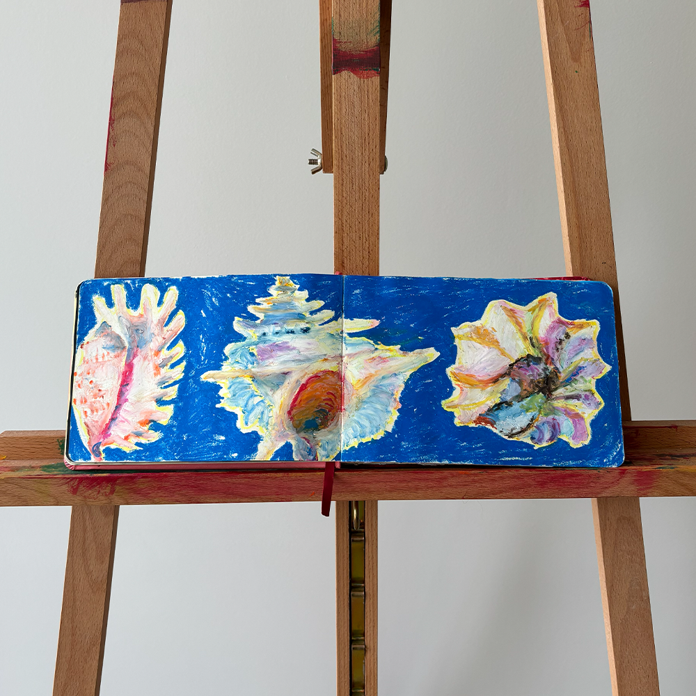 Livro aberto com pinturas de folhas e conchas, colocado em cavalete de madeira.