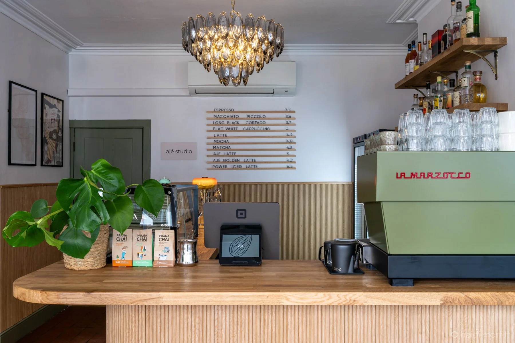 Elegant café counter with La Marzocco espresso machine, coffee menu, liquor shelves, hanging glasses, chandelier, and potted monstera plant
