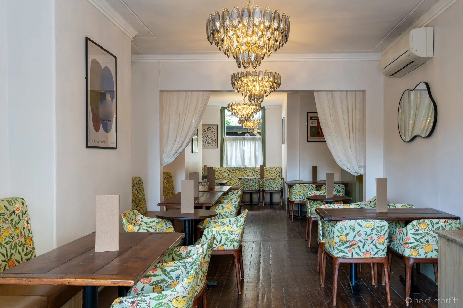 Elegant restaurant dining room with floral upholstered chairs, wooden tables, crystal chandeliers, white curtains, and wall art