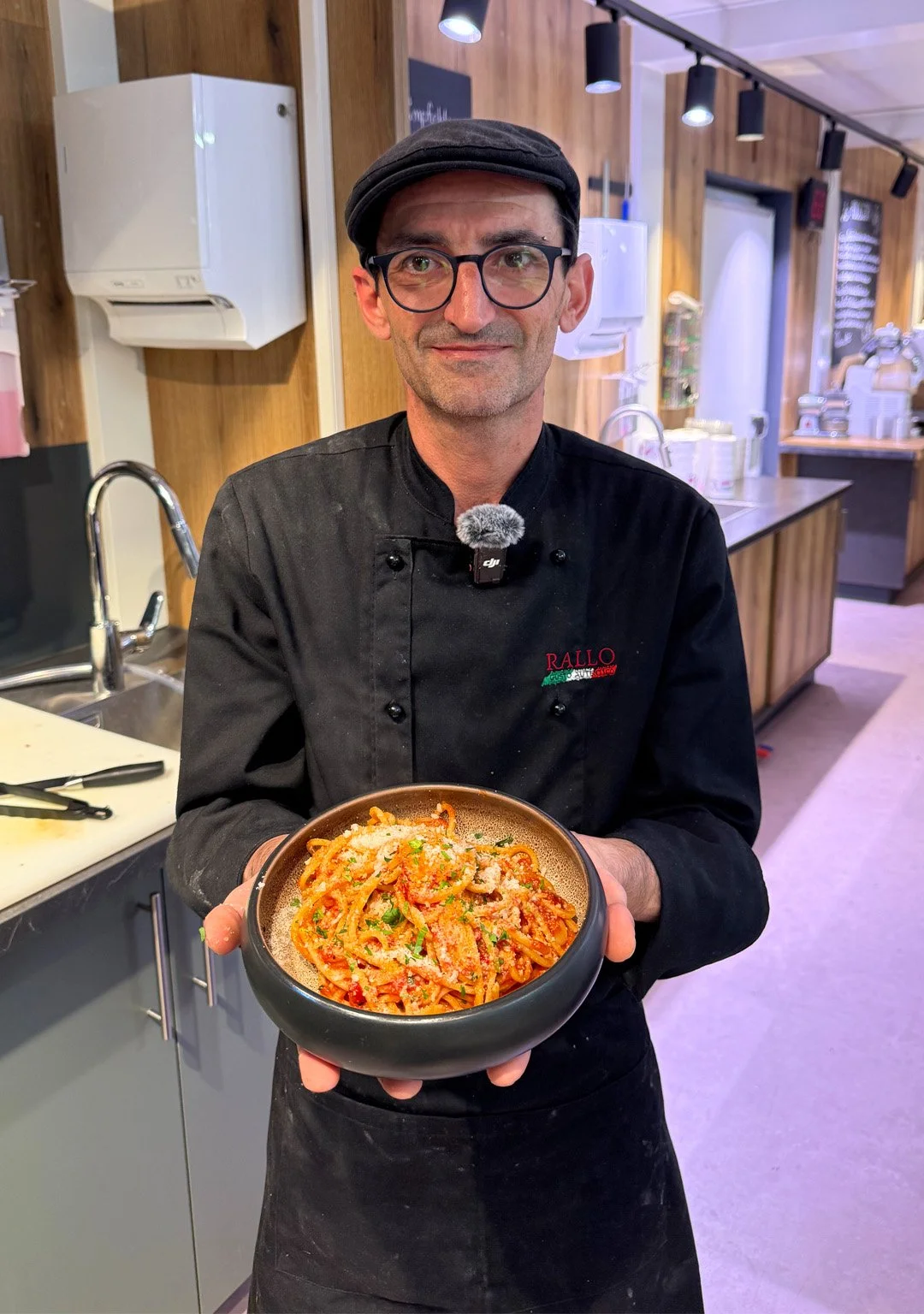 Ein Mann trägt eine schwarze Kochjacke mit rotem und grünem Logo, hält eine Schüssel mit Pasta, wahrscheinlich Spaghetti mit Tomatensauce, in einem Restaurantküche.