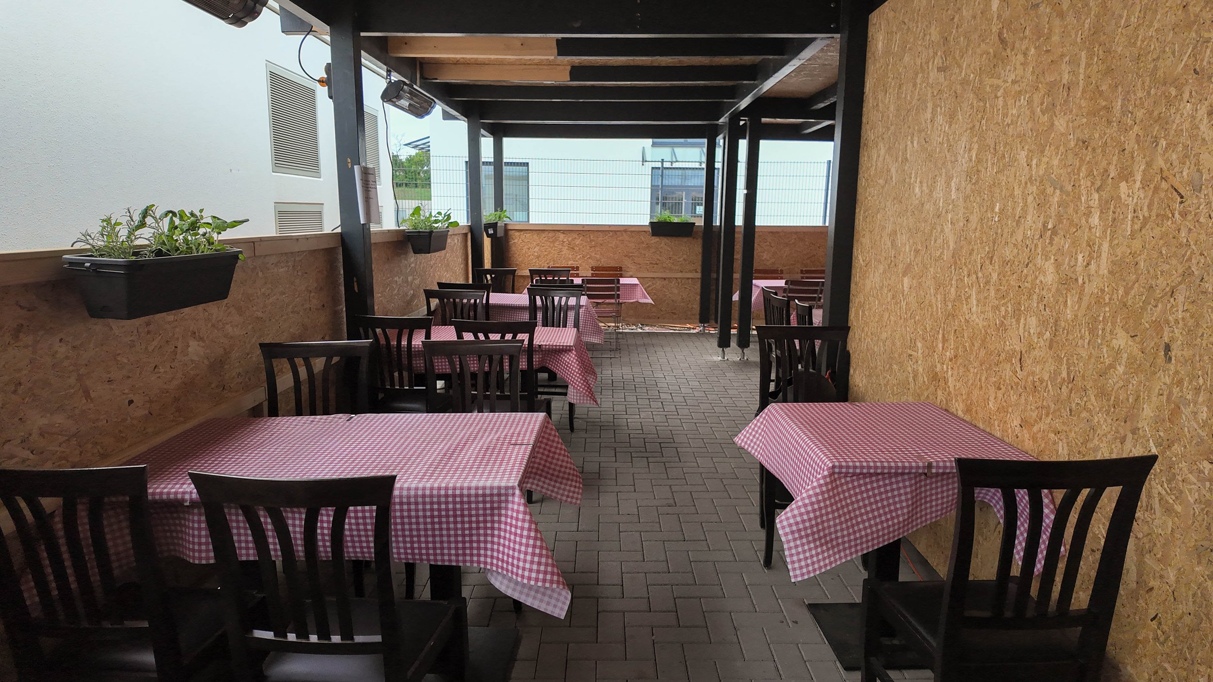 Blick auf einen überdachten Außenbereich eines Restaurants mit Tischen, die mit roten und weißen karierten Tischdecken bedeckt sind, mit schwarzen Stühlen, Pflanzen an der Wand und einem Netzzaun im Hintergrund.