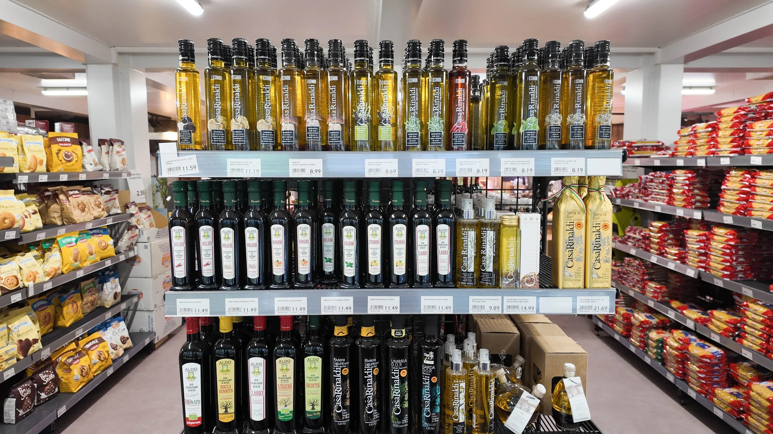 Regal mit verschiedenen Olivenölflaschen in einem Supermarkt, umgeben von Snack-Produkten auf den Regalen.
