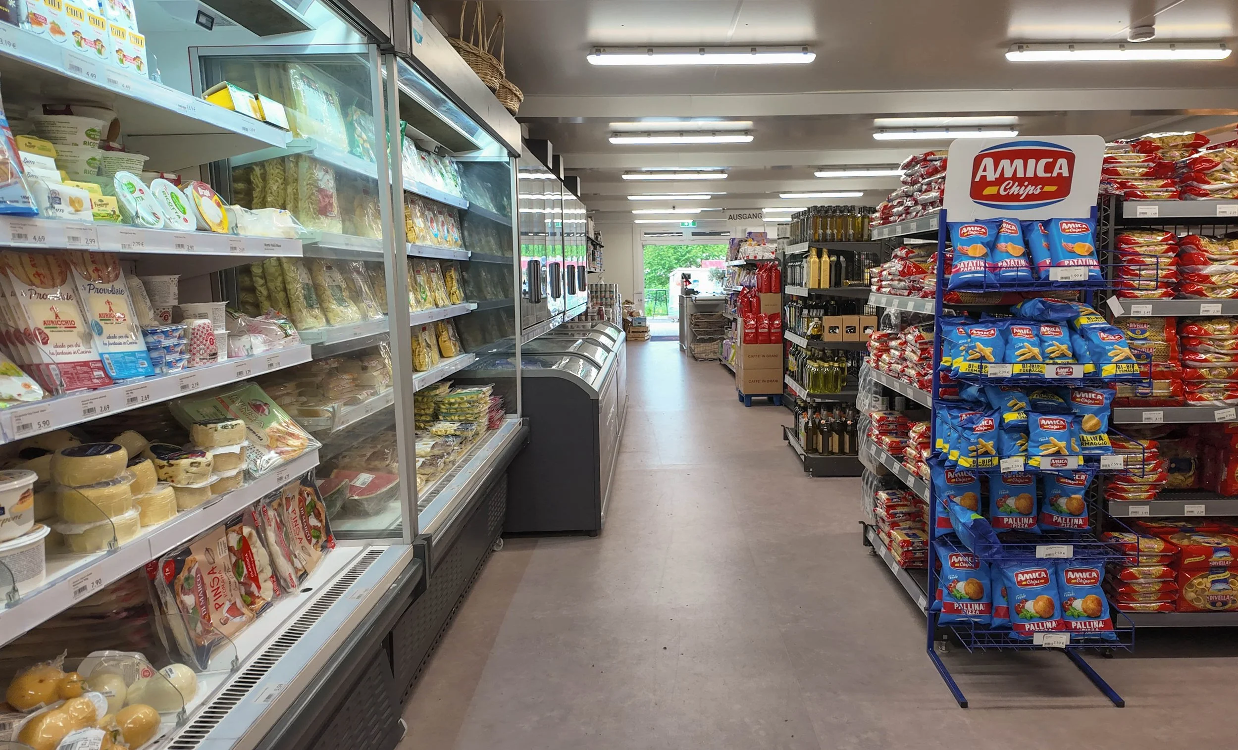 Aisle in a supermarket with shelves filled with food products, including cheese, pasta, and snack chips.