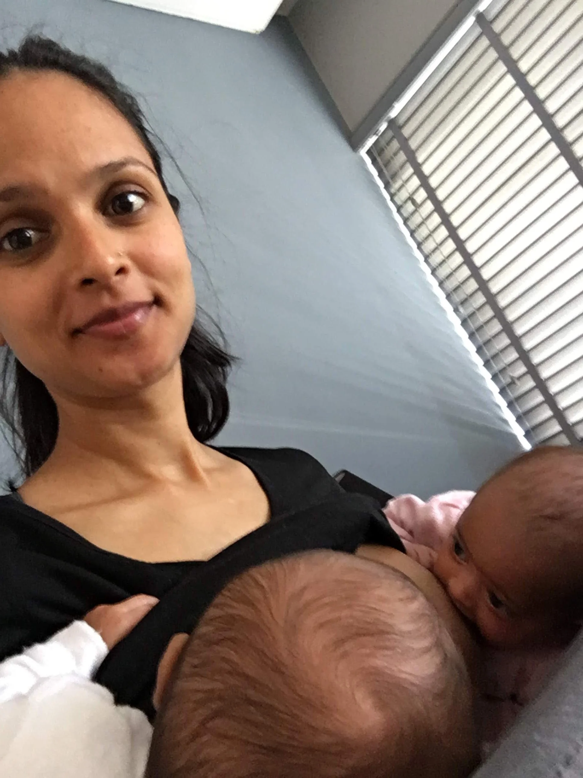 A woman with dark hair and a black top is smiling while holding two infants, one with light brown hair and the other with a bald head, close to her chest. Breastfeeding mother of twins is feeling proud of her perseverance in daily life.