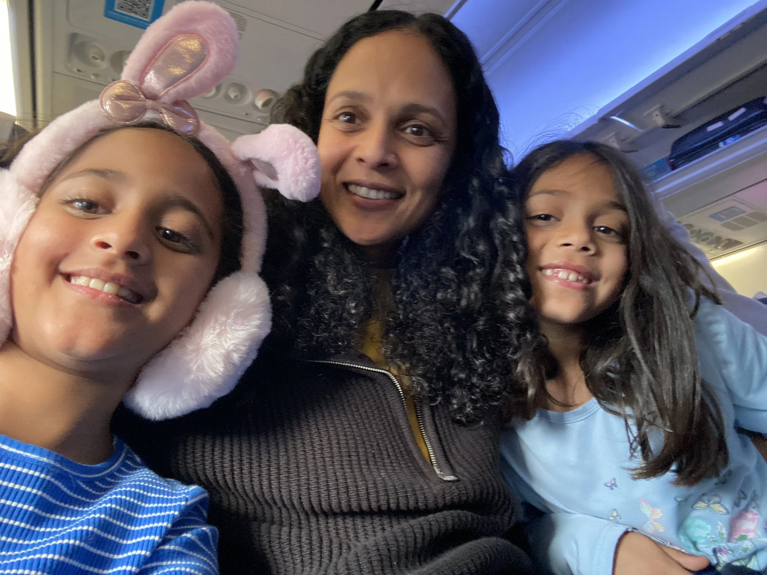 A woman smiling with two young girls, one wearing pink bunny ears, sitting inside an airplane. Family therapist with her children using ear phones to help regulate in noisy environments.