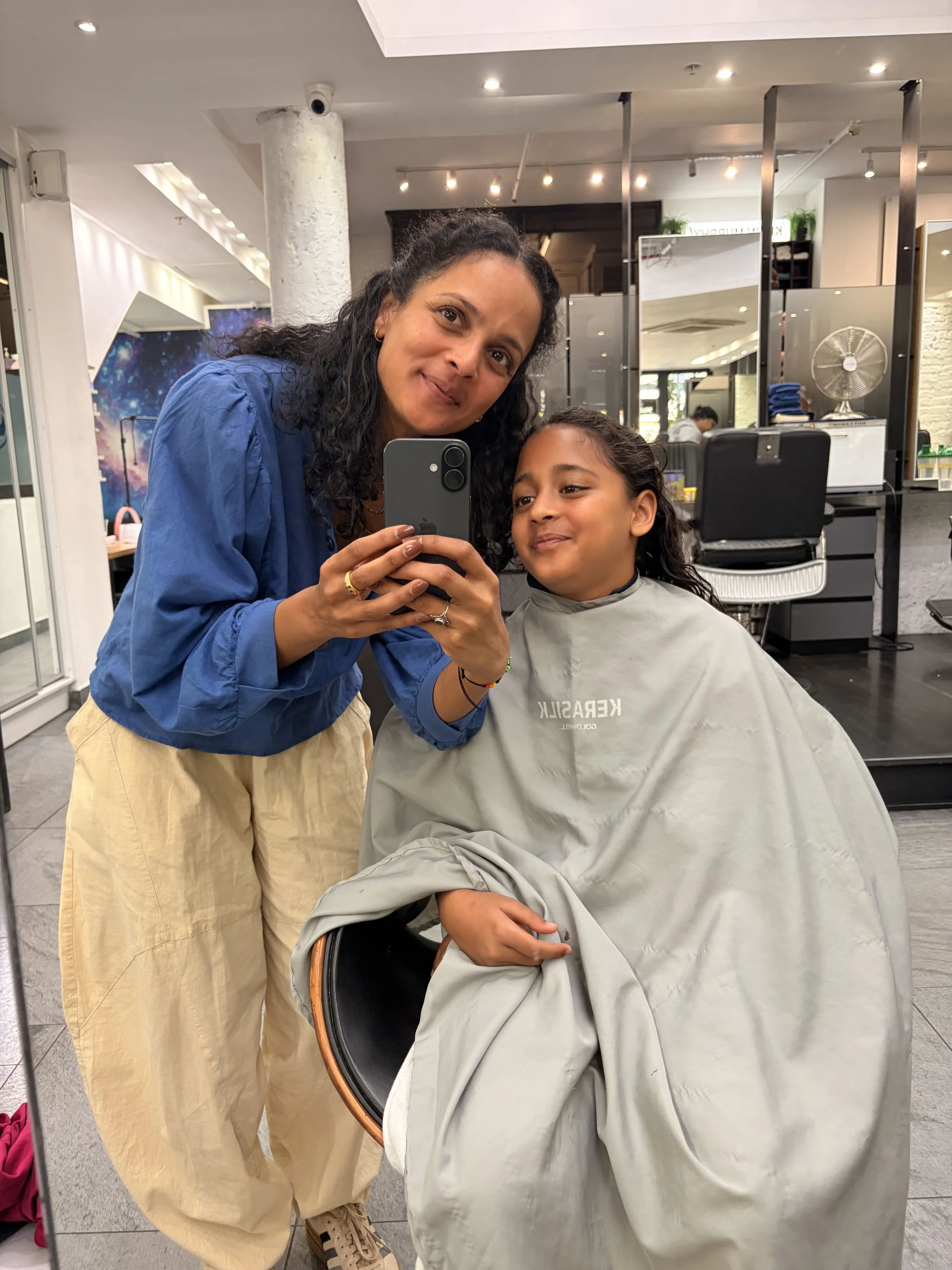A woman and a girl taking a selfie in a salon. The woman has dark, curly hair and is smiling at the camera. The girl has dark hair tied back and is sitting in a salon chair with a cape. The connection with child and mother is unmatched.