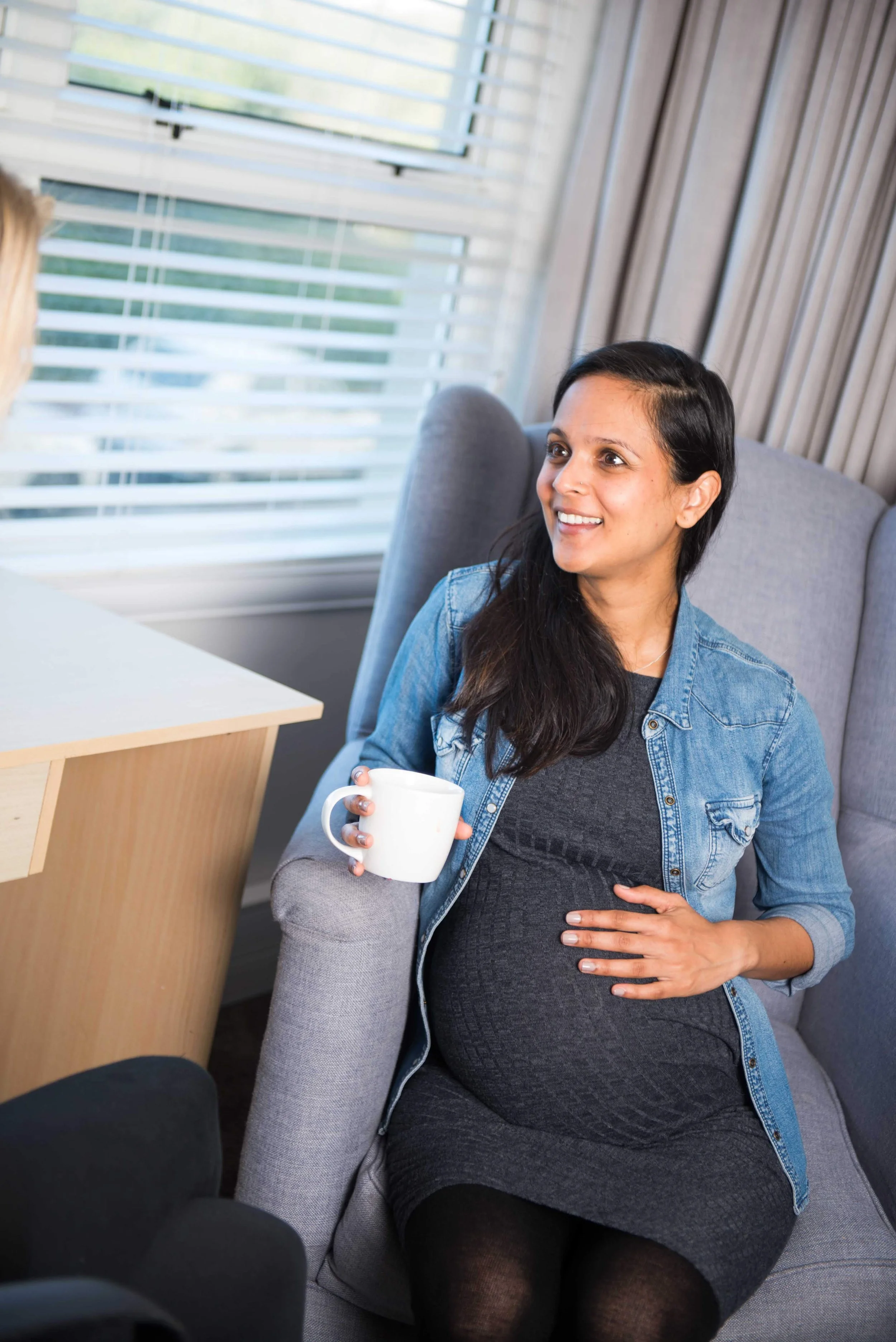 A pregnant woman with long dark hair, wearing a gray dress and a denim jacket, sitting on a gray armchair and holding a white mug, smiling, in a room with horizontal window blinds. Receiving support for post natal depression and parental burnout.