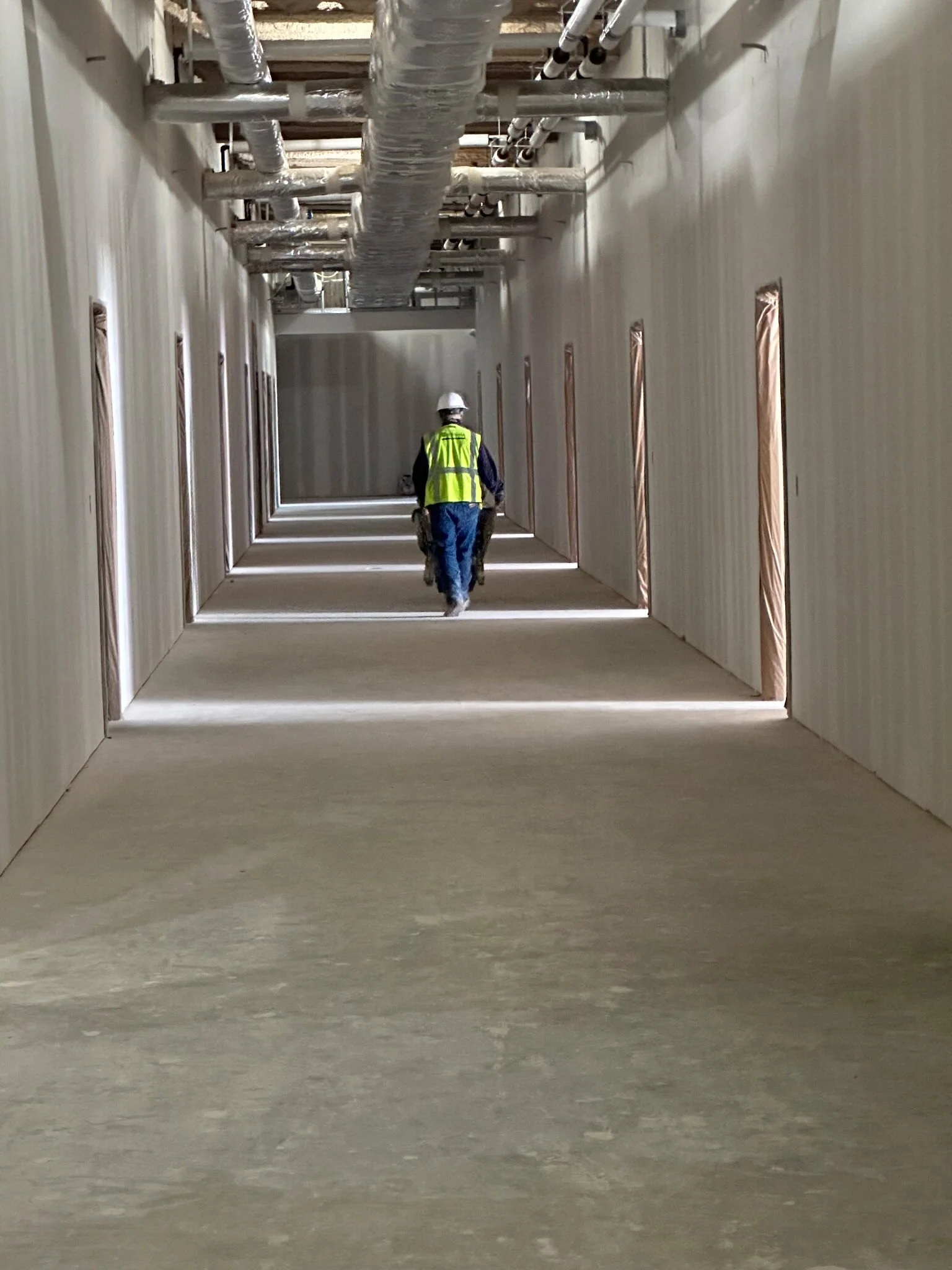 Construction worker walking down unfinished hallway with exposed ductwork on ceiling and framed wall openings on sides.
