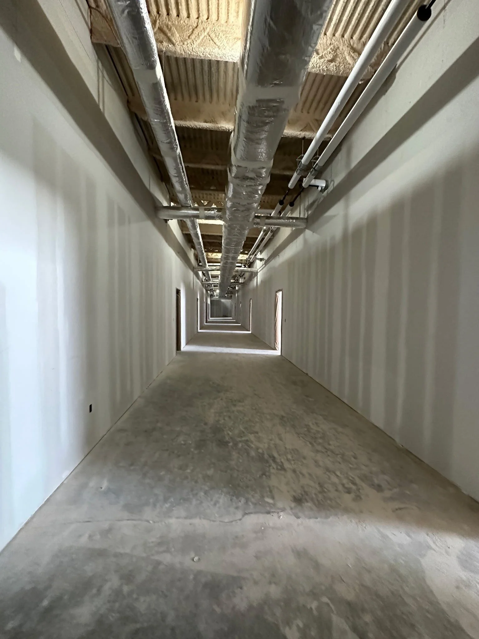 Long hallway under construction, with unfinished flooring, white walls, and exposed ceiling with ductwork and piping.