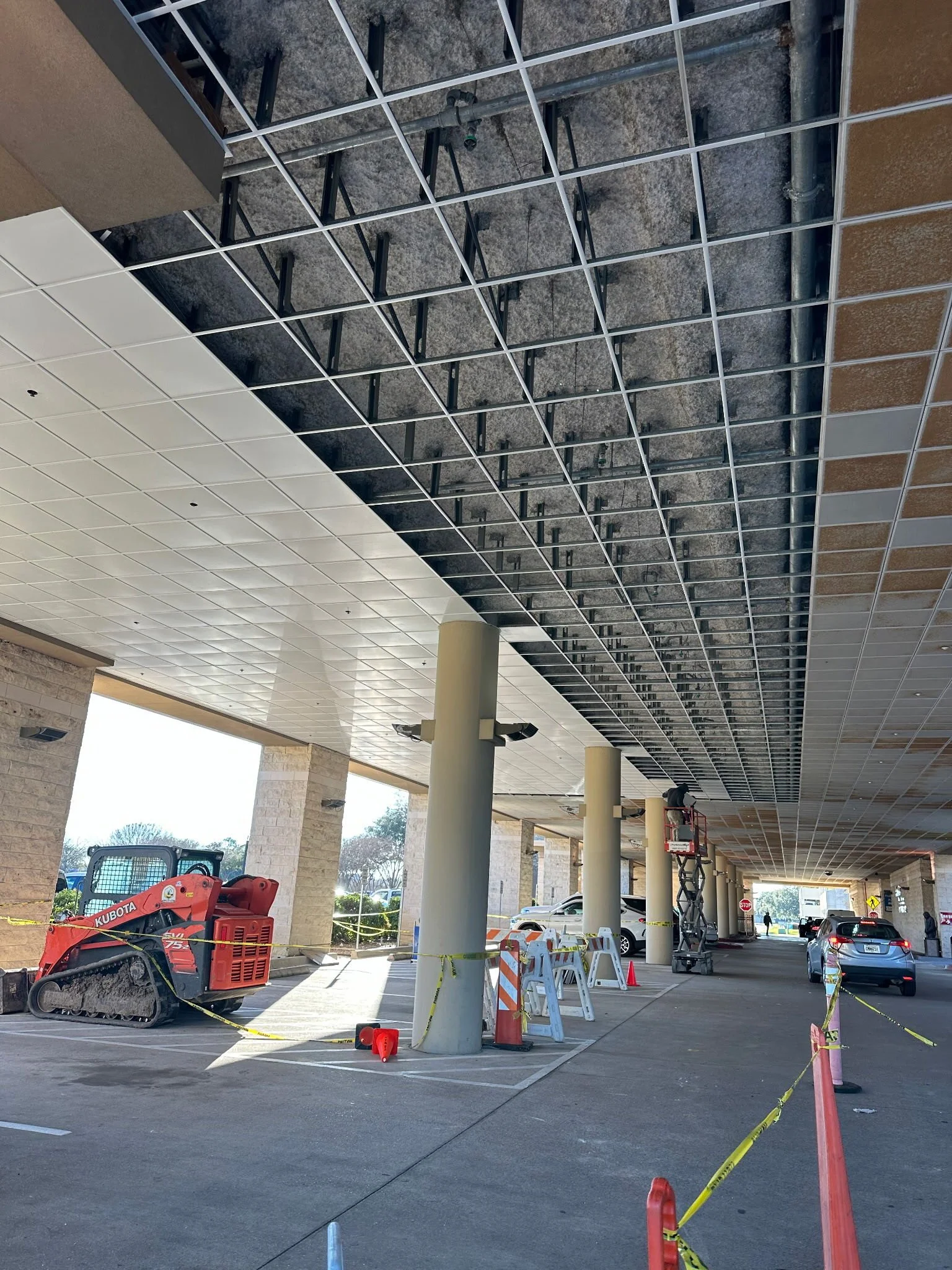 Construction workers installing ceiling panels in a parking garage, with equipment and safety barriers in place.