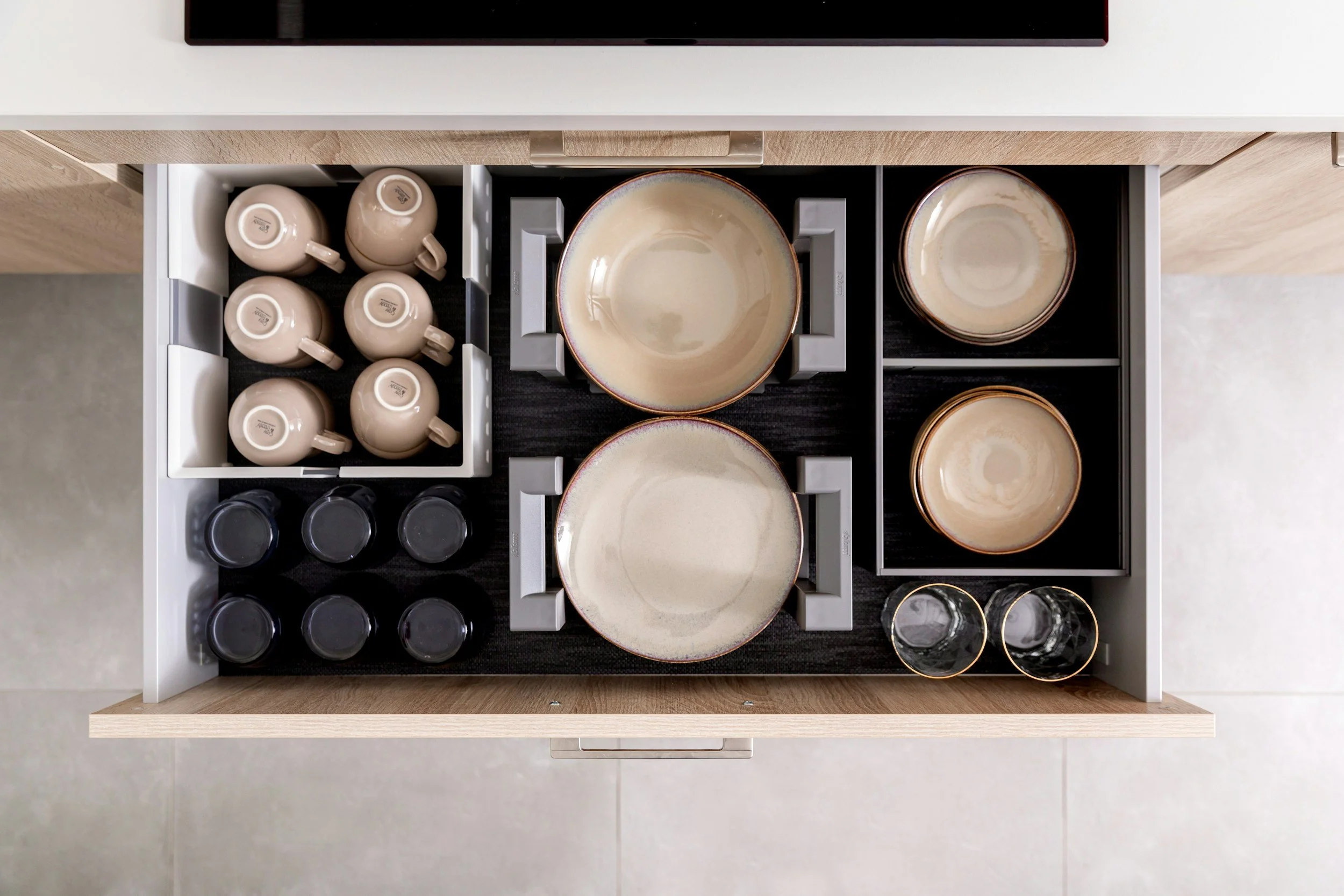 An open kitchen drawer containing beige cups and plates, black coasters, and clear glasses, organized neatly within individual compartments.