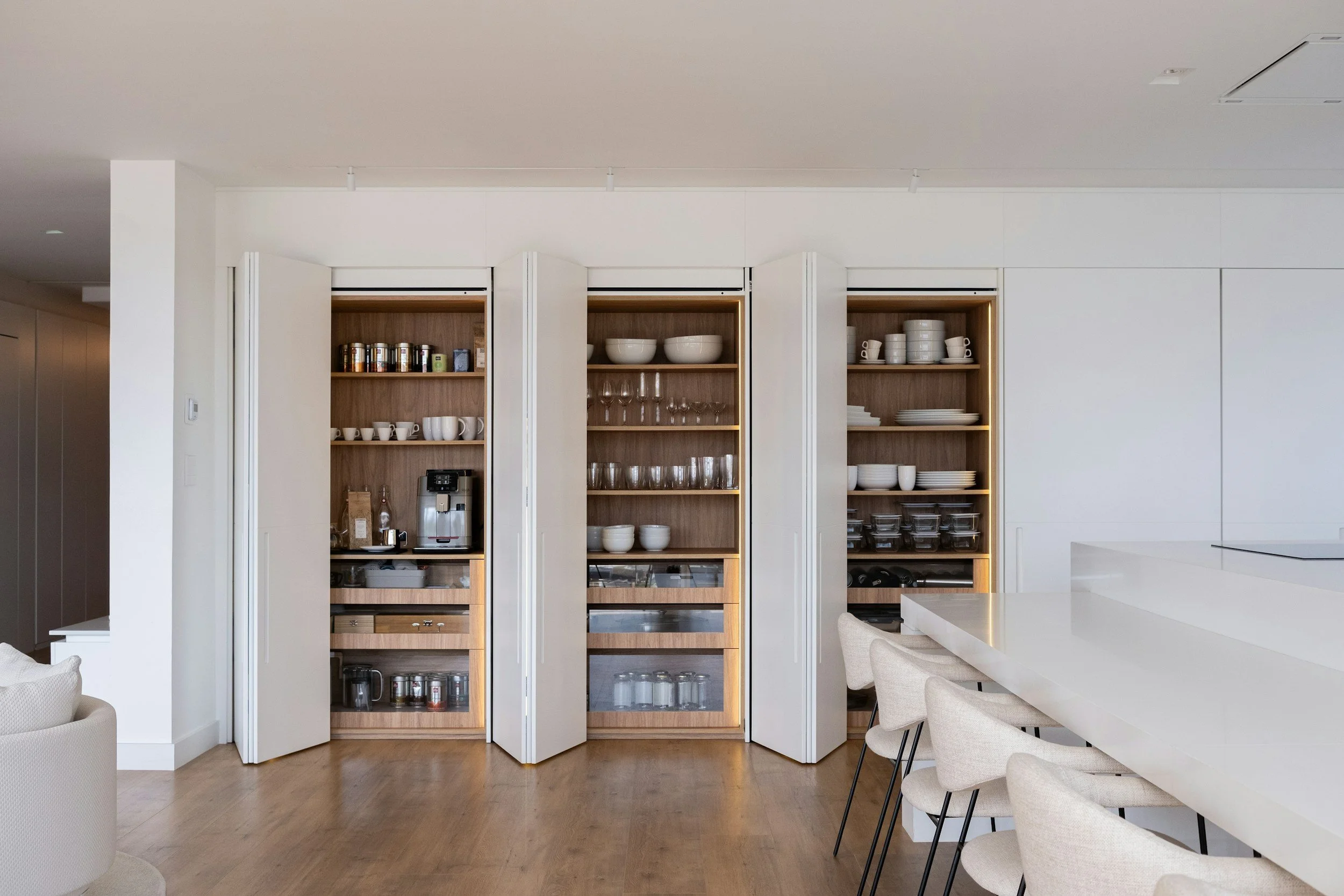 Open kitchen cabinets filled with white bowls, glasses, and cups, with a coffee machine on the countertop and a white dining table with beige chairs in a modern, minimalist kitchen.