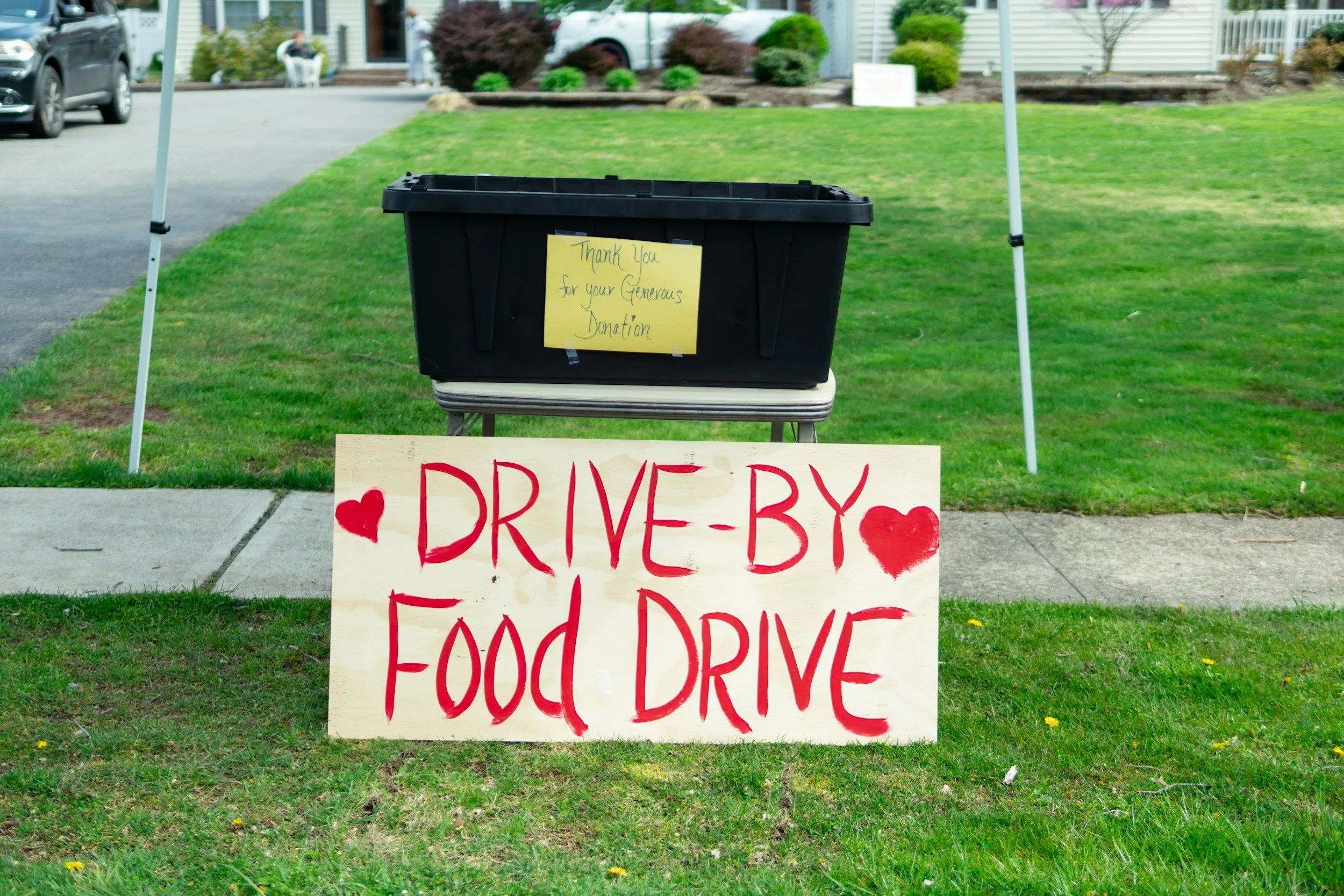 A black donation collection bin with a yellow sign that says, "Thank you for your generous donation." The bin is placed on a small table on a grassy lawn, with a large sign in front that reads, "Drive-by food drive" with red hearts on either side of the text. There are houses and cars in the background.