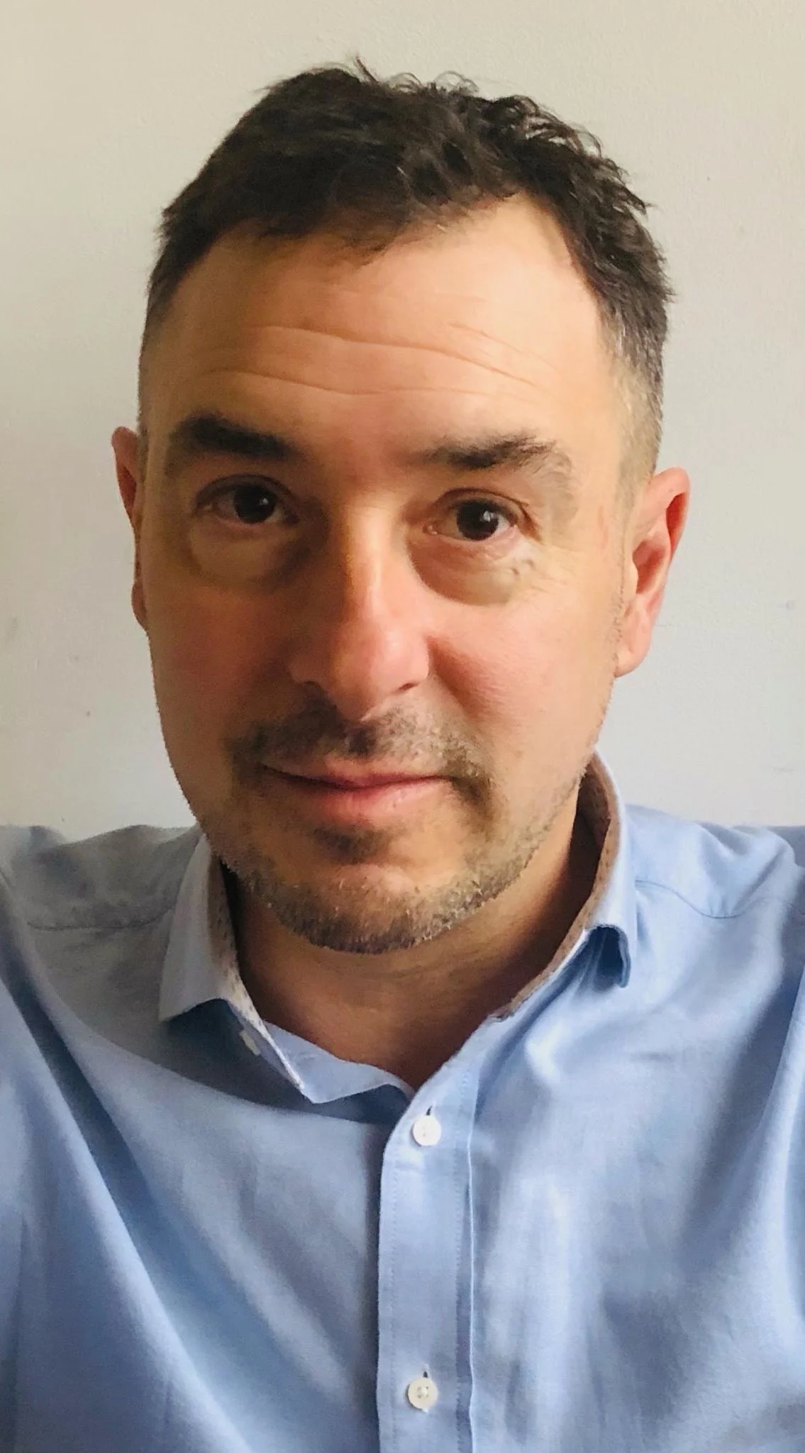 A middle-aged man with short, dark hair, facial stubble, and light skin, wearing a light blue collared shirt, looking directly at the camera, against a plain light-colored background.