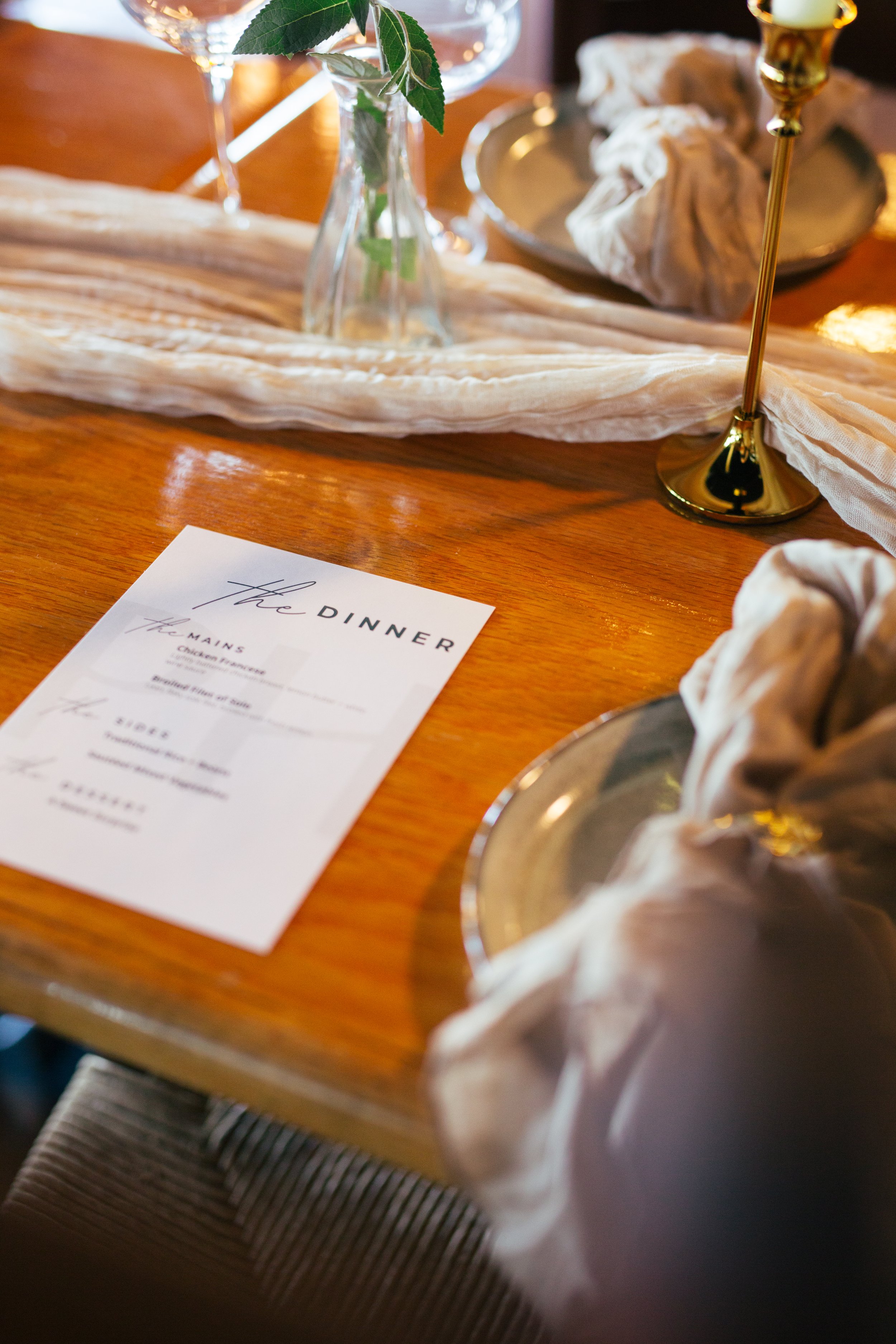A table setting with a dinner menu, a glass vase with green leaves, and gold candles, with a modern tablecloth and a wooden table surface.