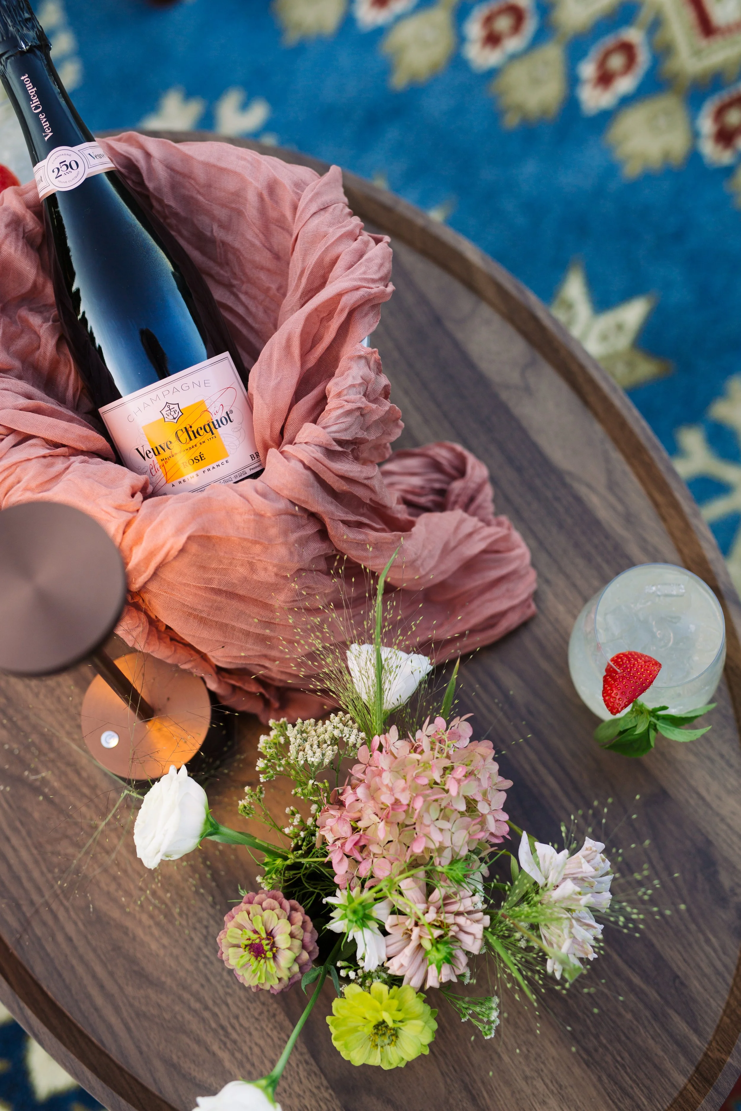 A bottle of Veuve Clicquot champagne placed in a pink cloth on a wooden table, surrounded by a floral arrangement with various white, pink, and yellow flowers, and a glass of sparkling water garnished with a strawberry.