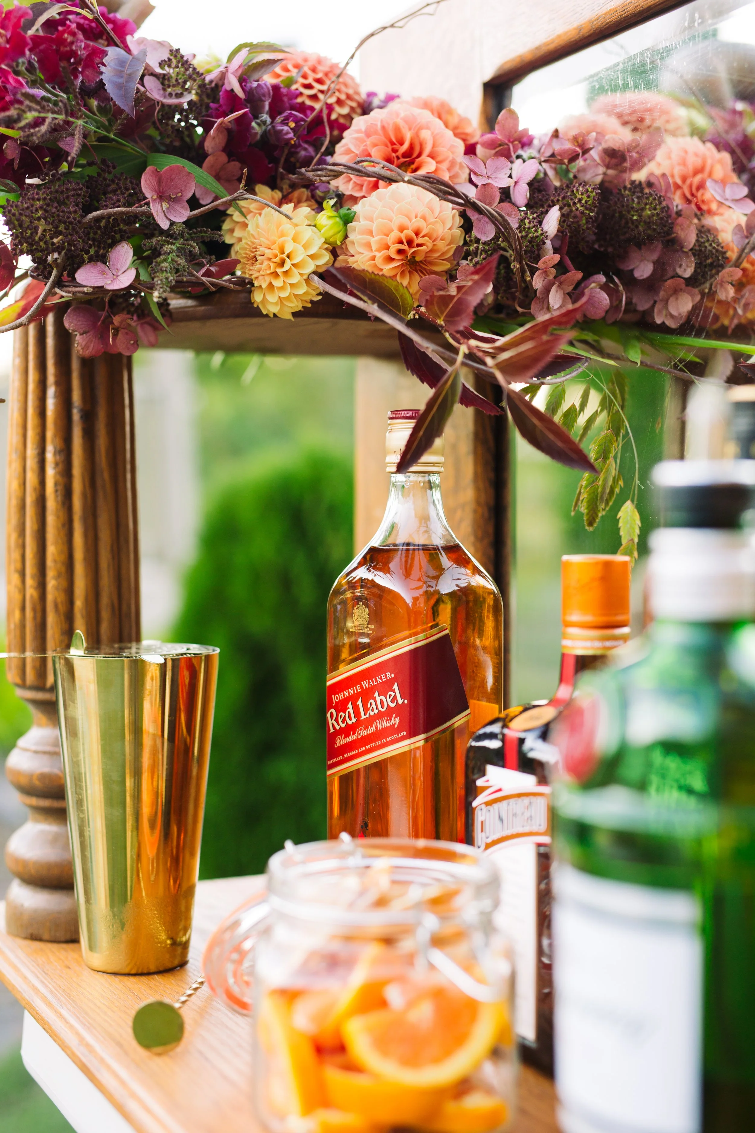 A close-up of a bar setup with a floral arrangement, a bottle of Johnnie Walker Red Label whiskey, and various drink ingredients on a wooden table.