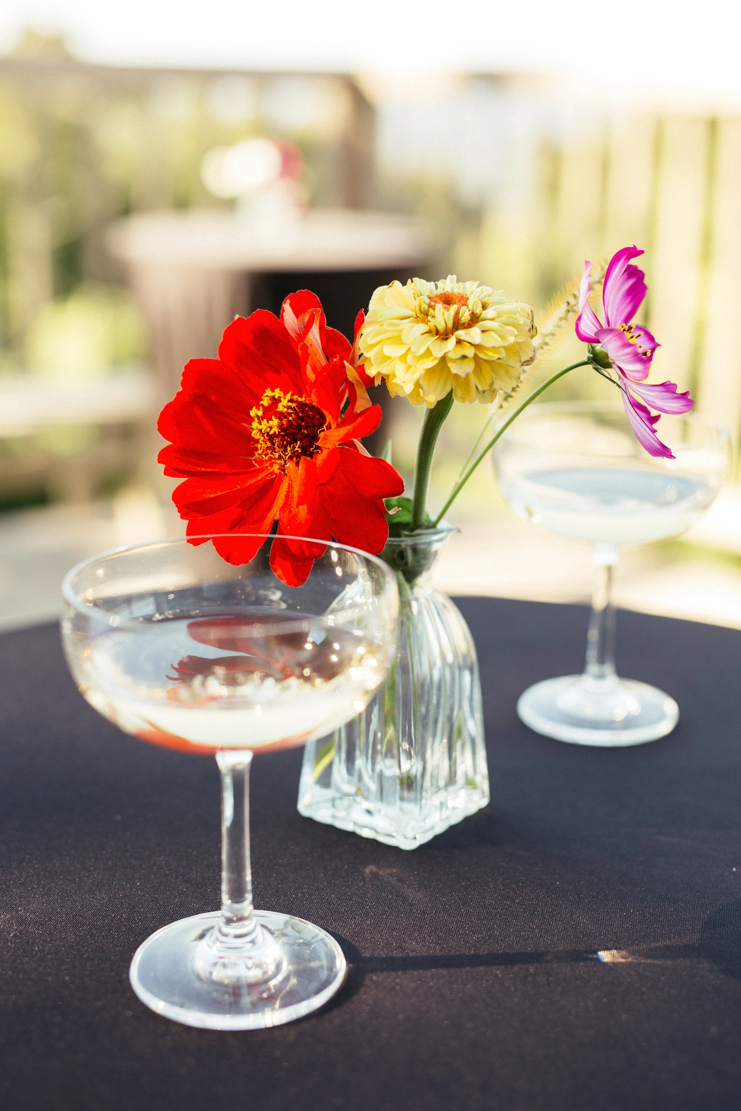 A clear glass vase with three colorful flowers—red, yellow, and pink—placed on a black tablecloth, with a glass of water and a blurred outdoor background.