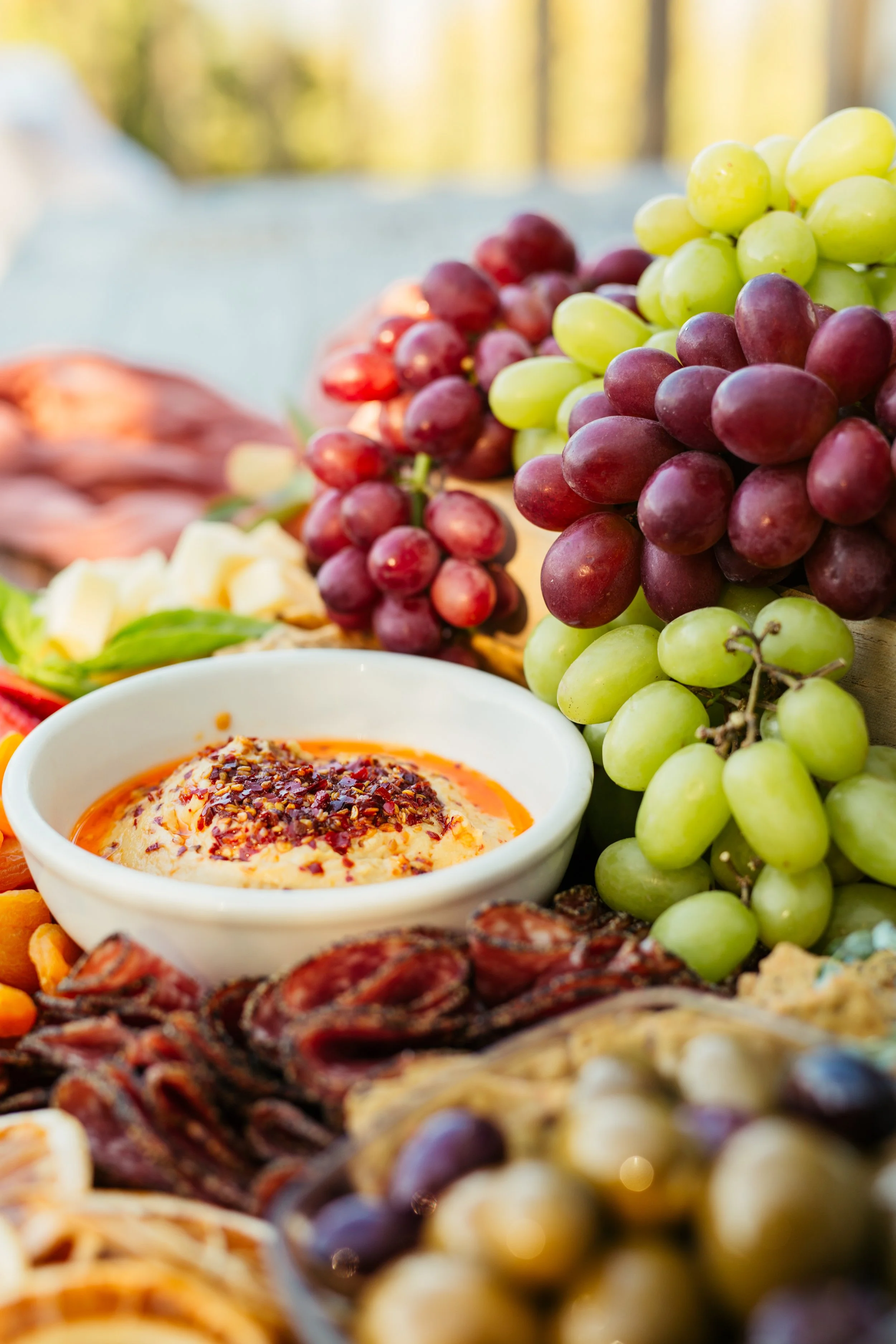 Close-up of a cheese dish with red pepper flakes, surrounded by green and purple grapes, and other assorted foods.