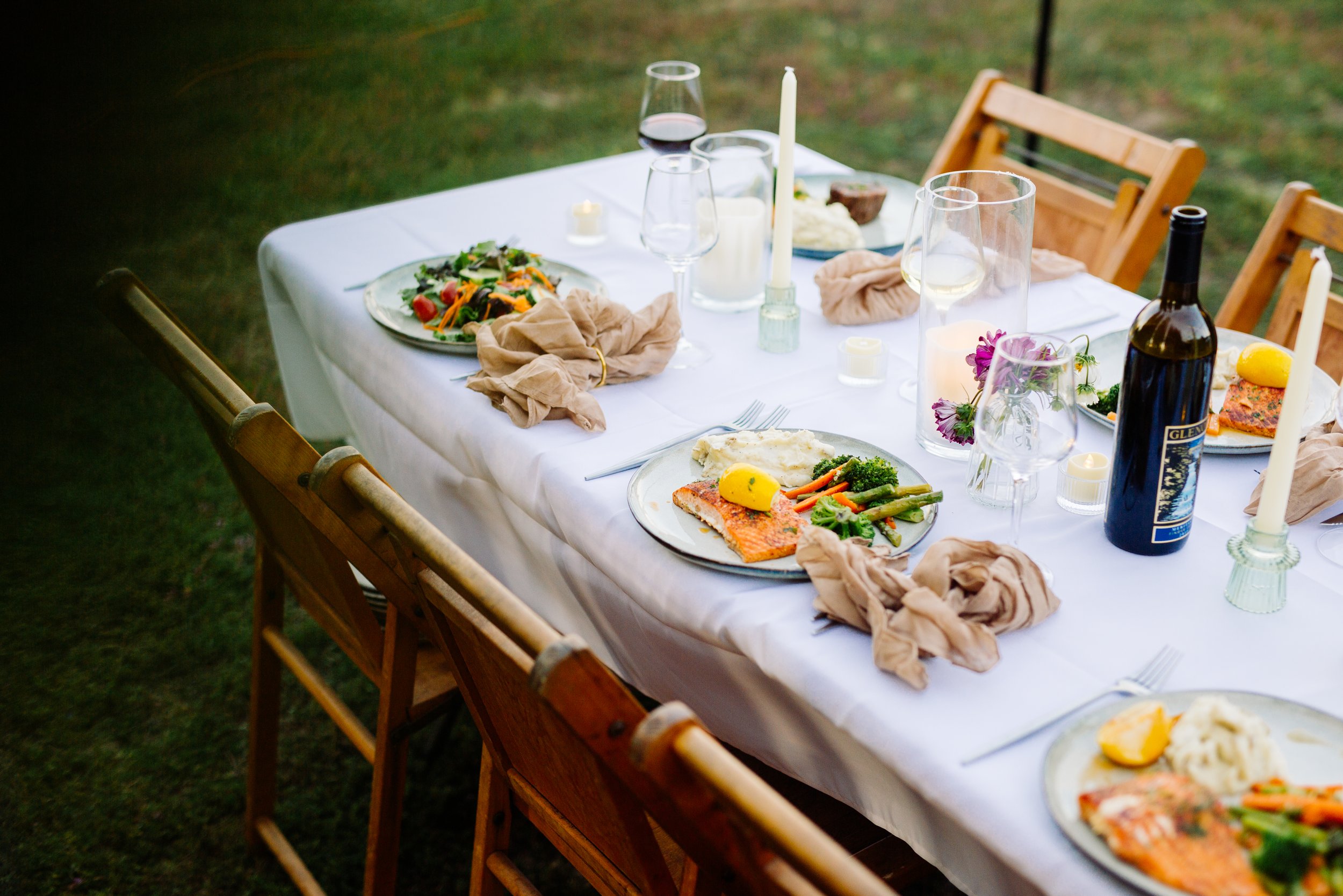 A table set outdoors with plates of food, wine glasses, candles, and a wine bottle.