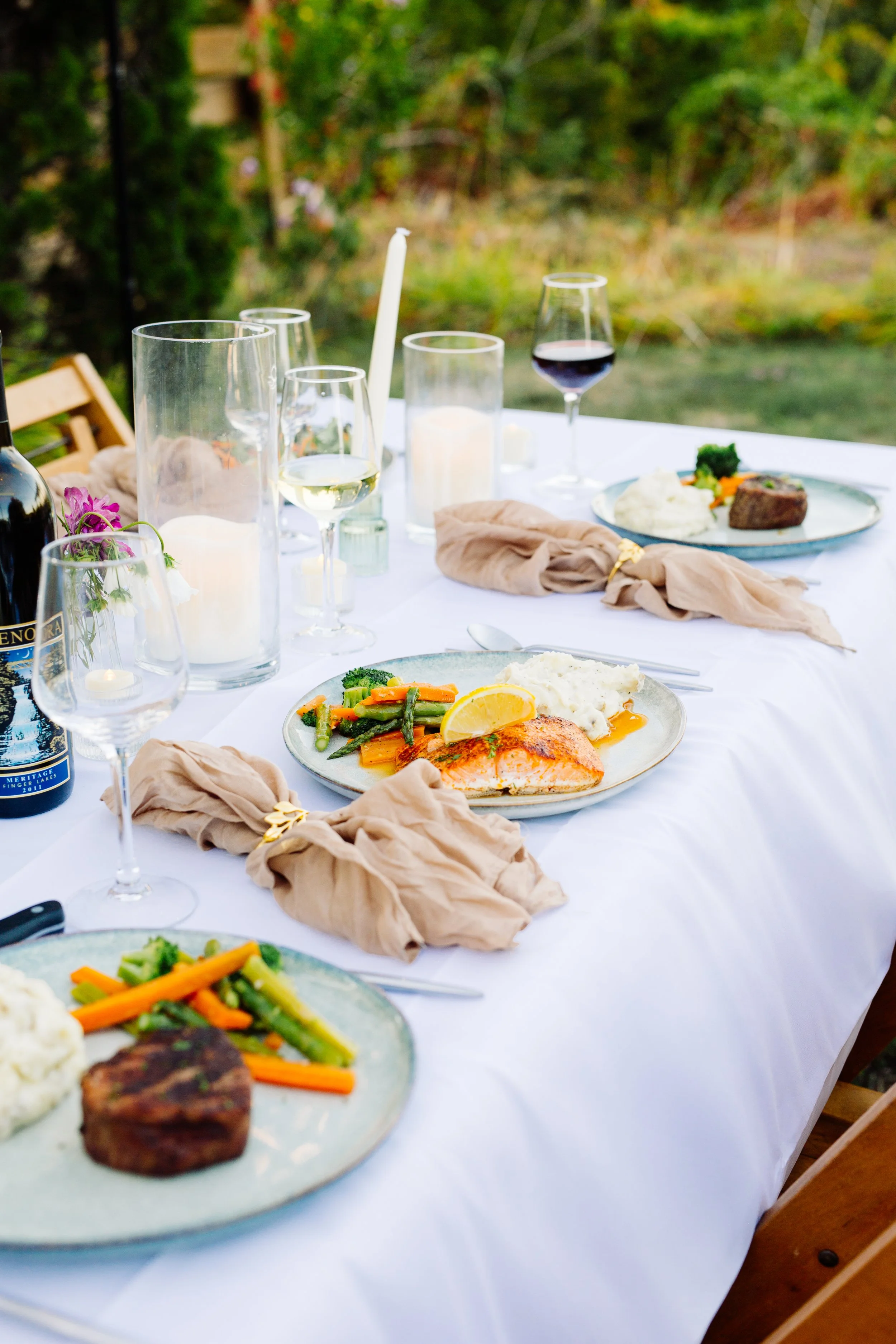 A table set with plates of food, wine glasses, a candle, and a napkin, outdoors with greenery in the background.