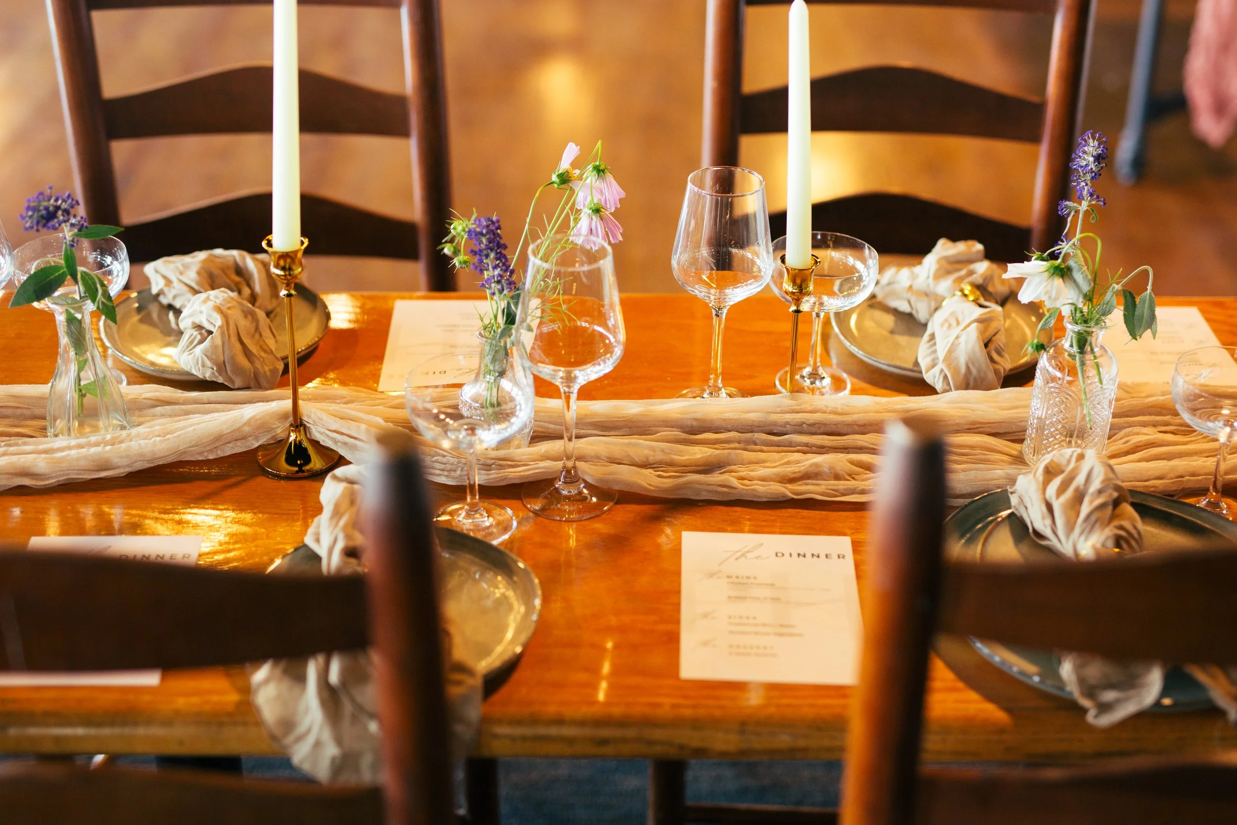 A dining table set with candles, flowers in small vases, plates, wine glasses, and cloth napkins with a menu card underneath.