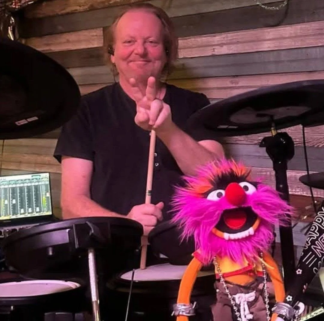 A man with light skin, smiling, holding drumsticks behind electronic drums. A colorful puppet with pink and orange fur, large eyes, and an open mouth is in front of the drums.