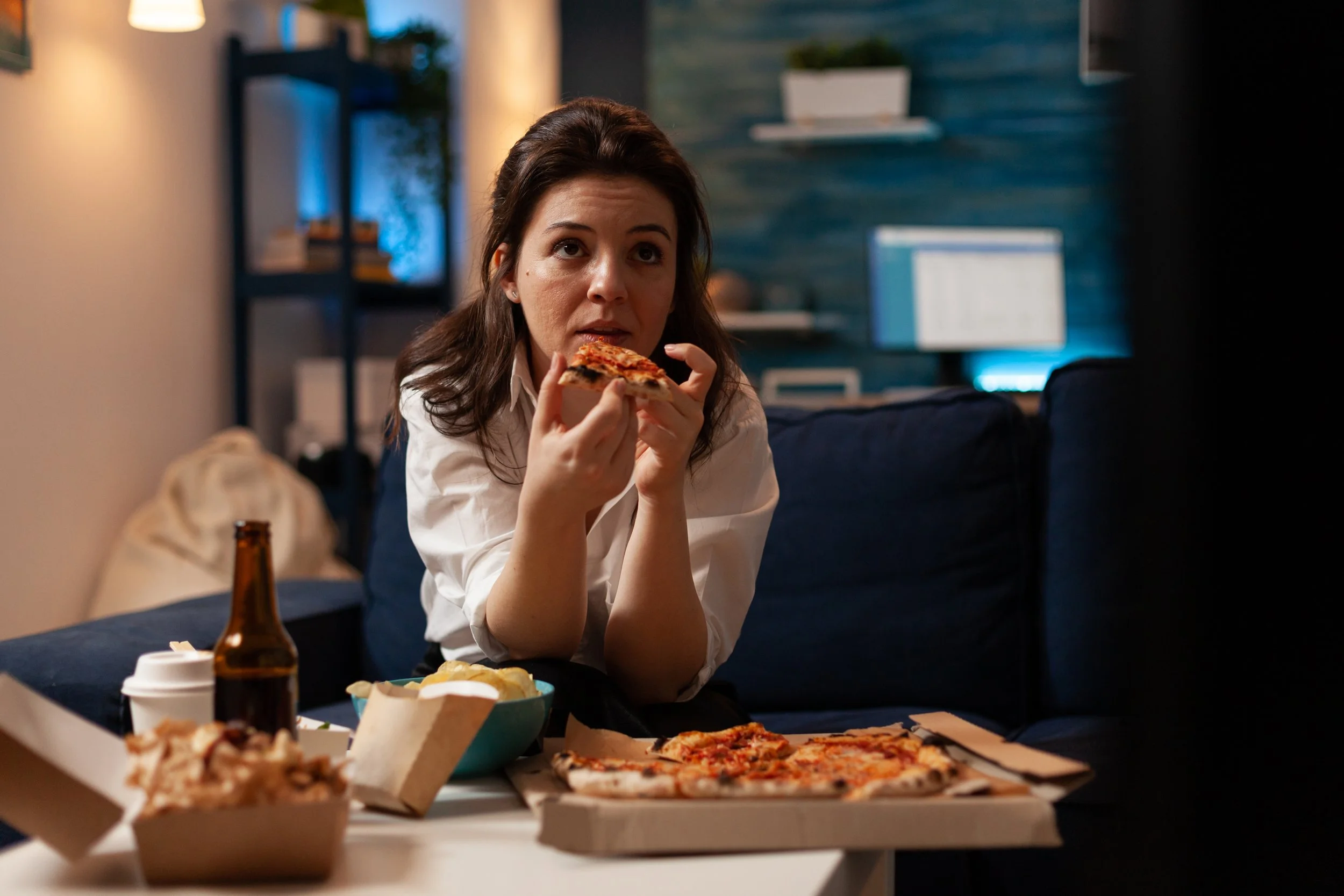 A woman with dark brown hair eating a slice of pizza at a dining table. There are various food items including pizza and snacks on the table, along with a brown beer bottle and a white takeout coffee cup.