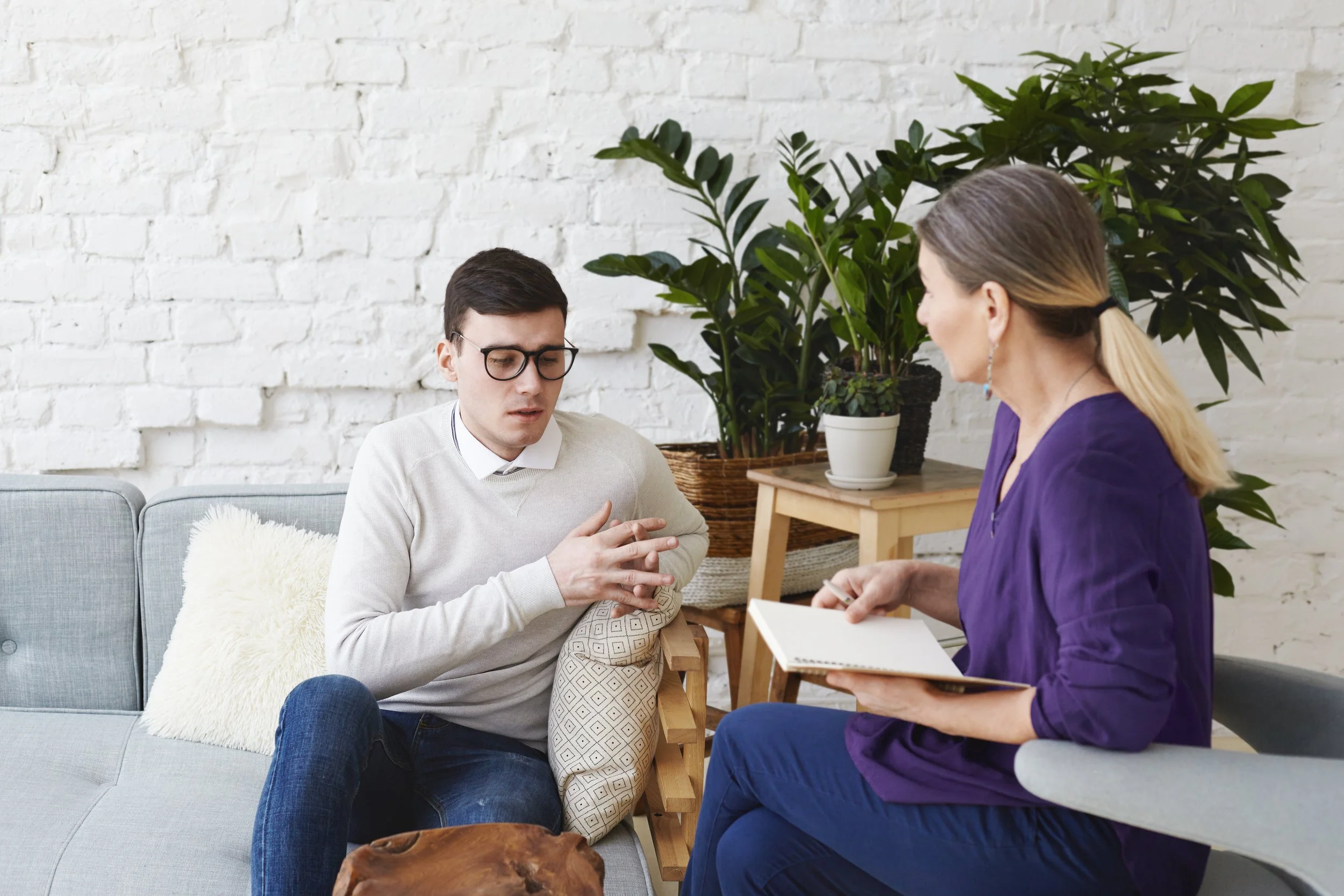 A young man with glasses and a woman with long hair in a ponytail engaged in a conversation in a cozy room with white brick walls and large green plants.