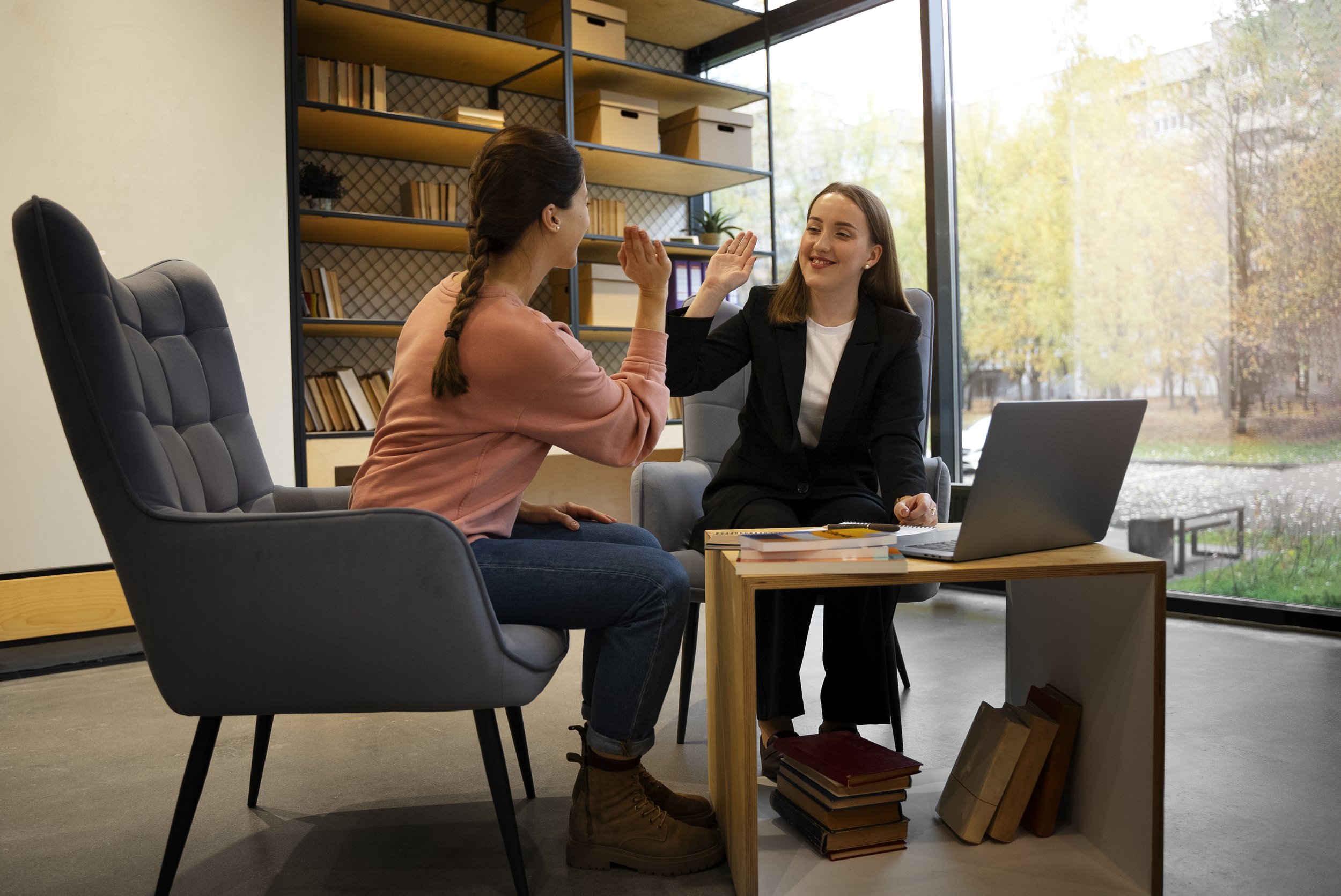 Two women sitting at a desk in a modern office, giving each other a high five, with a laptop and books nearby, large window revealing trees outside.
