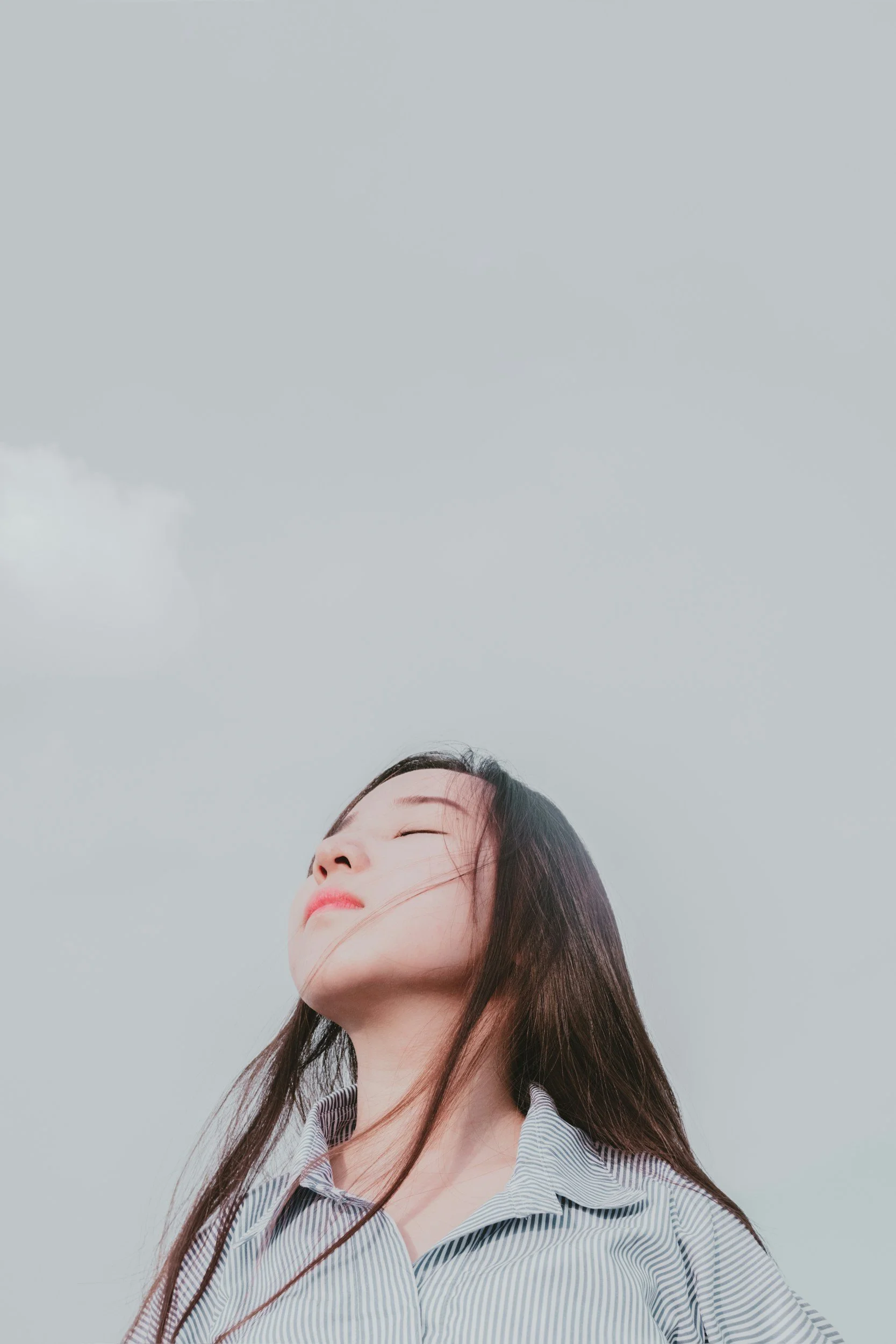 A woman with long dark hair and a striped shirt standing outdoors against a cloudy sky, with her eyes closed and face relaxed.