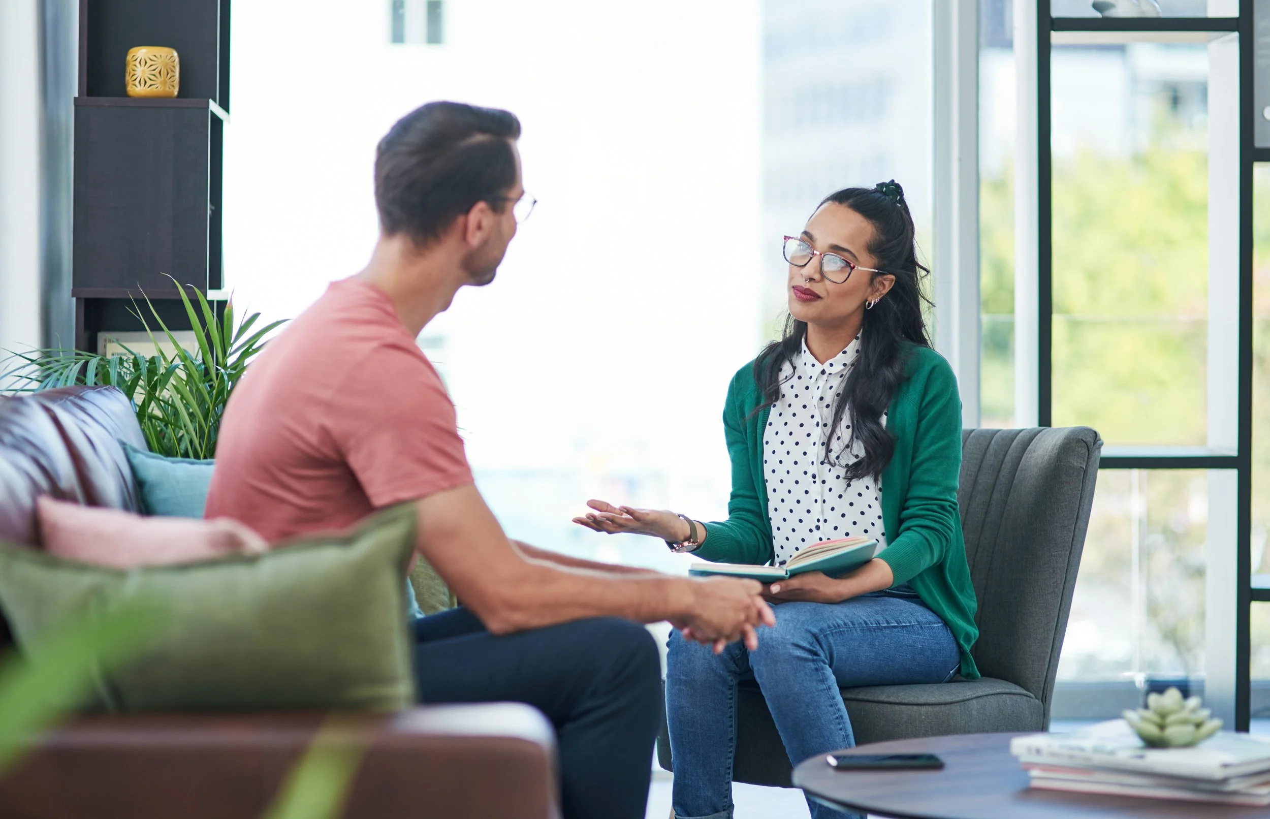 A woman with glasses, a polka dot shirt, and a green cardigan talking to a man with glasses and a pink shirt in a bright, modern room with large windows and indoor plants.