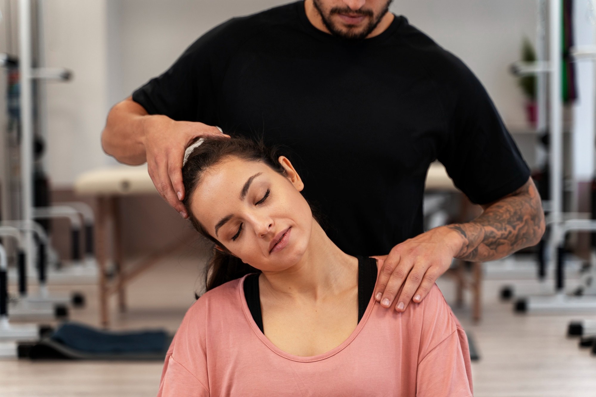 A chiropractor wearing a black t-shirt adjusting a woman's neck in a clinic. The woman is seated wearing a pink shirt with her eyes closed.