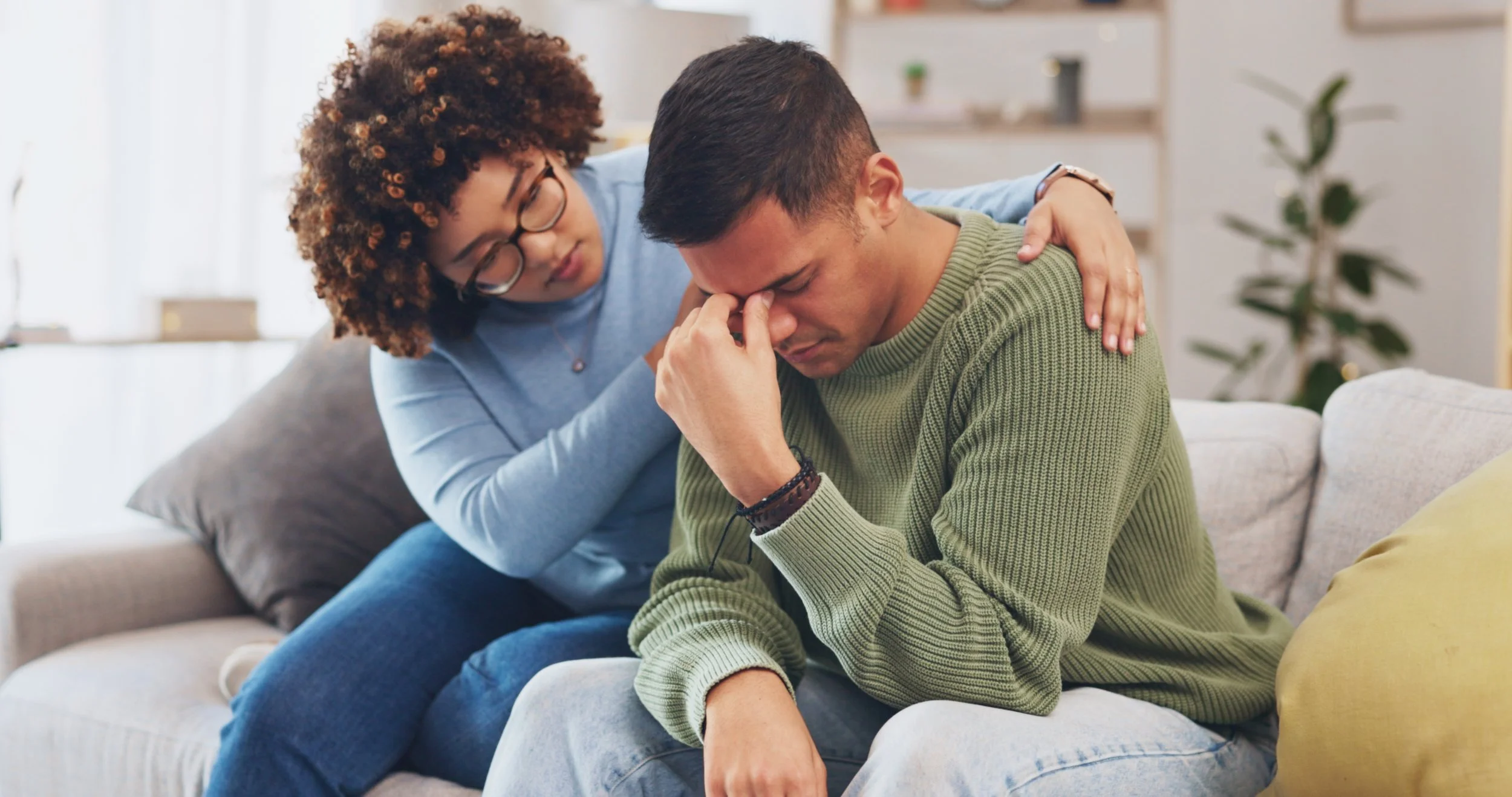 A woman is comforting a distressed man sitting on a couch, with her hand on his shoulder and his head bowed.