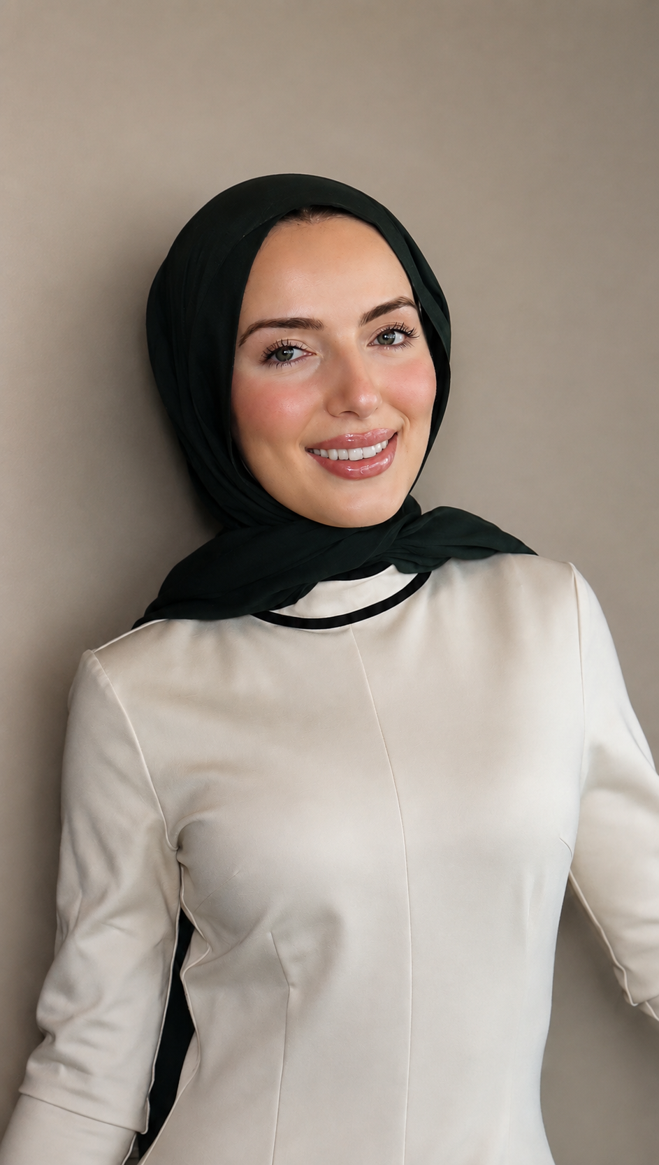 A smiling woman wearing a black headscarf and a cream-colored outfit, standing against a neutral background.